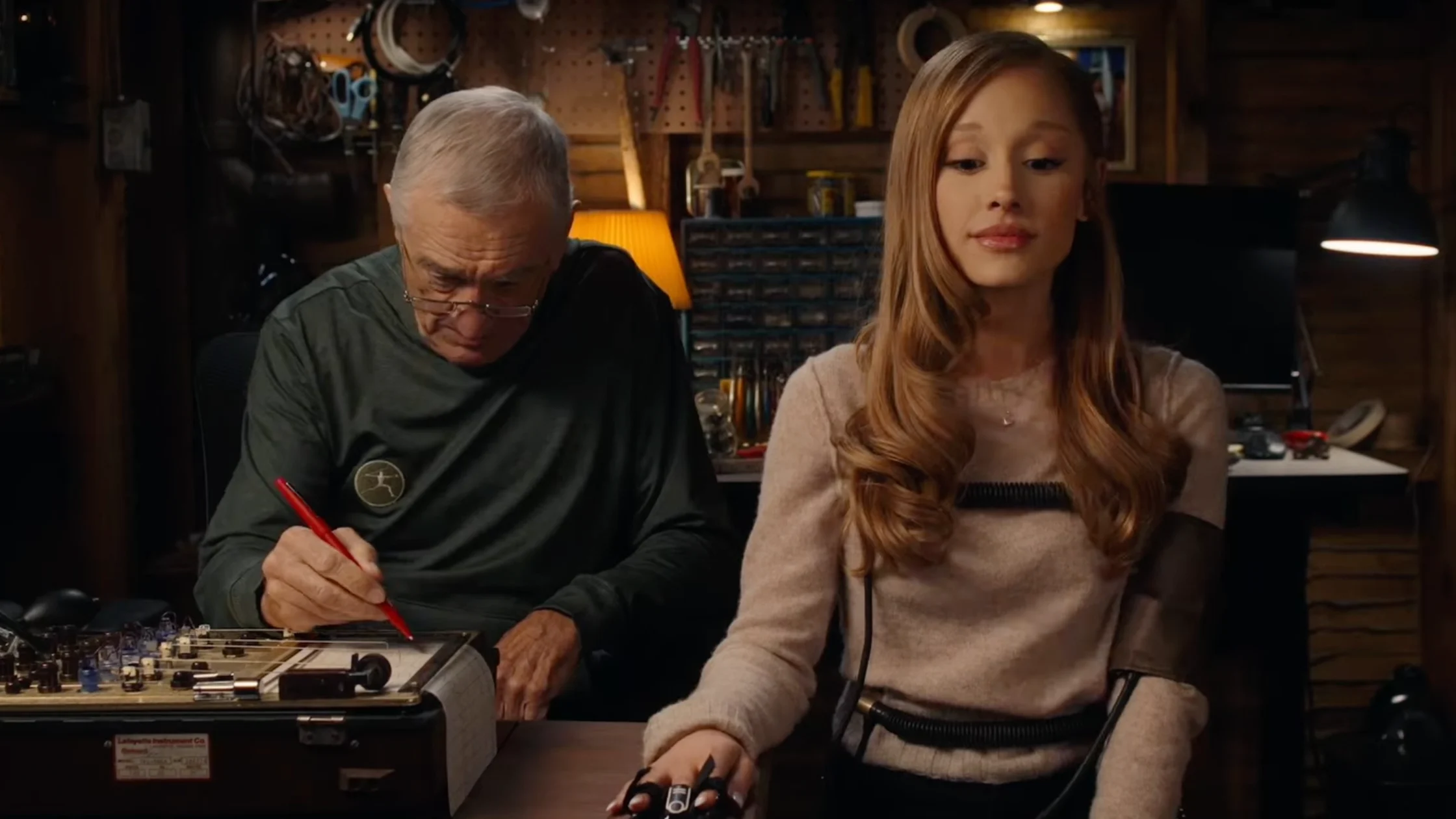 Older man with glasses and gray hair writes on an electronics project at a workbench while a young woman sits beside him in a tool-filled workshop.