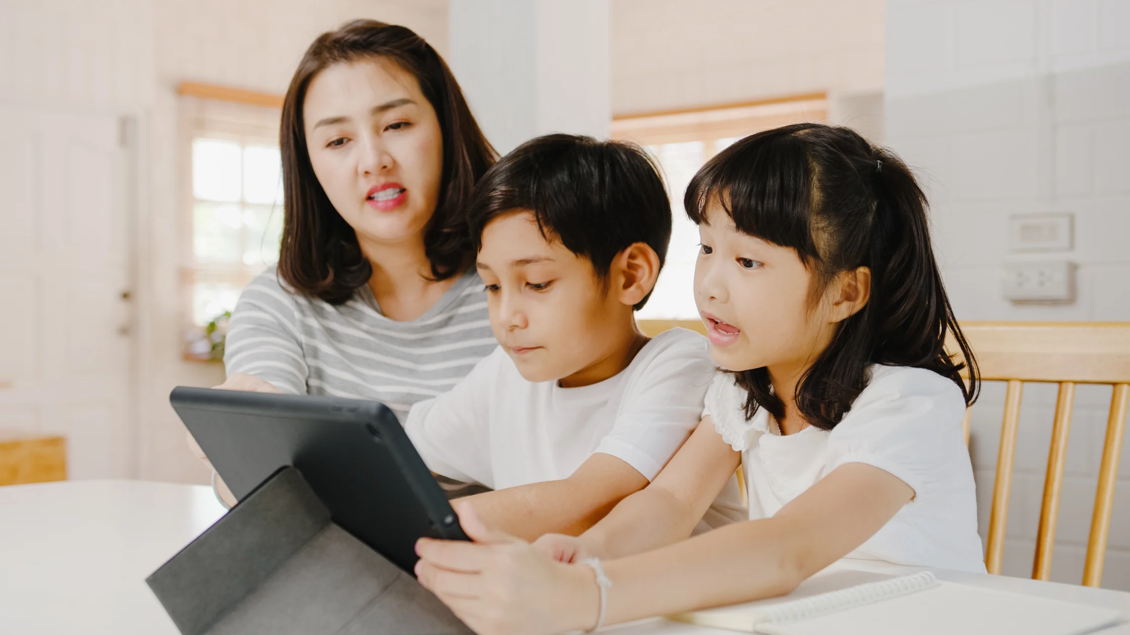 Mother and two children sit at a table, focused on a tablet screen together.