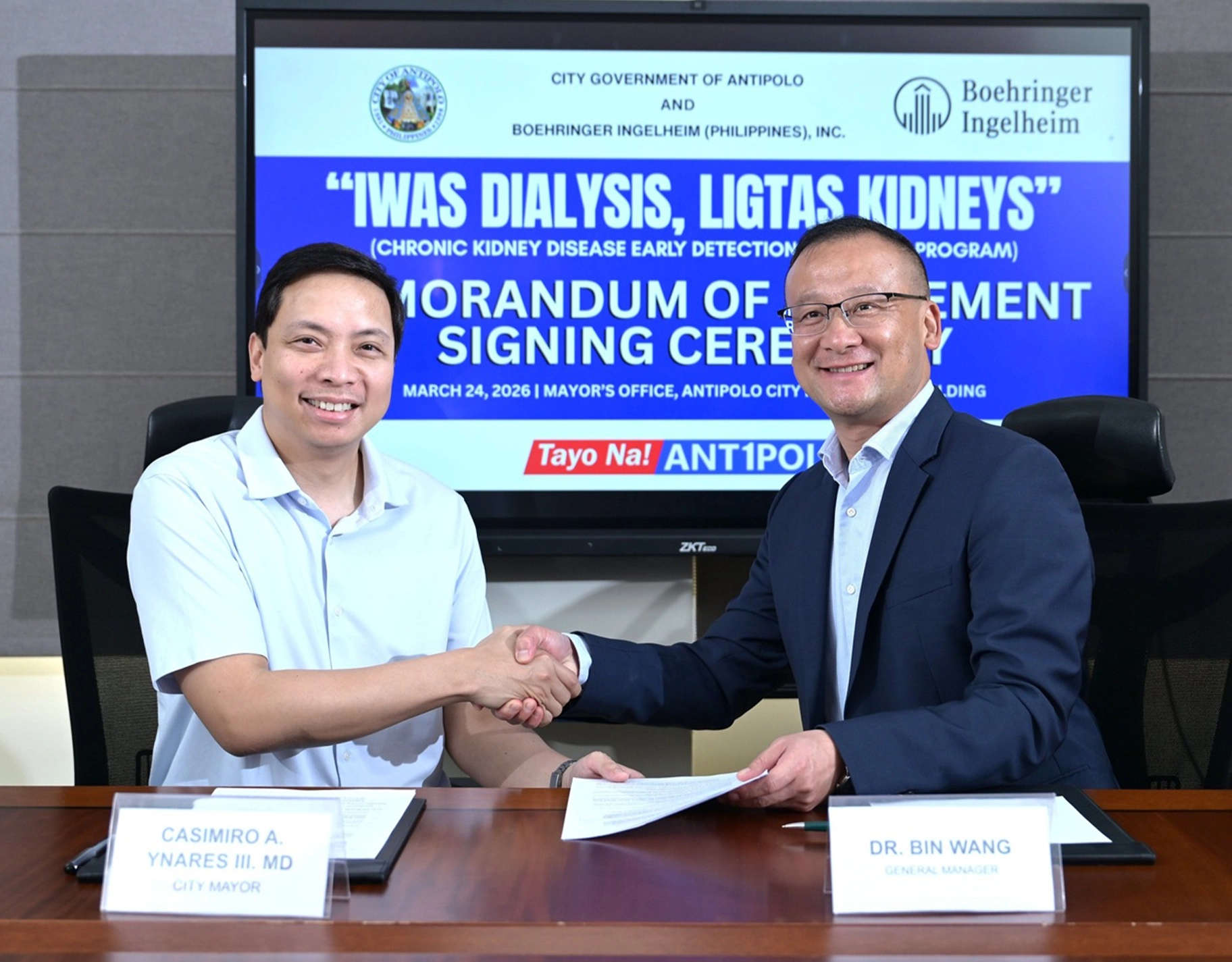 Two men shake hands across a conference table during a signing ceremony, with a large blue banner reading Memorandum of Understanding behind them.