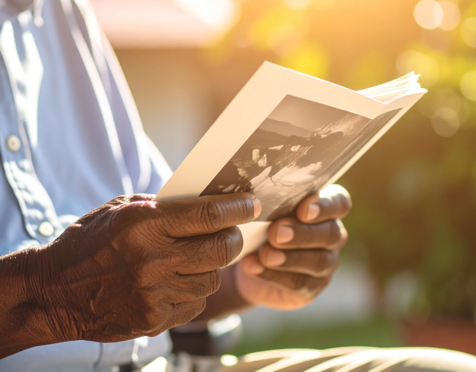 Close-up of dark-skinned hands holding an open photo album outdoors in warm sunlight.
