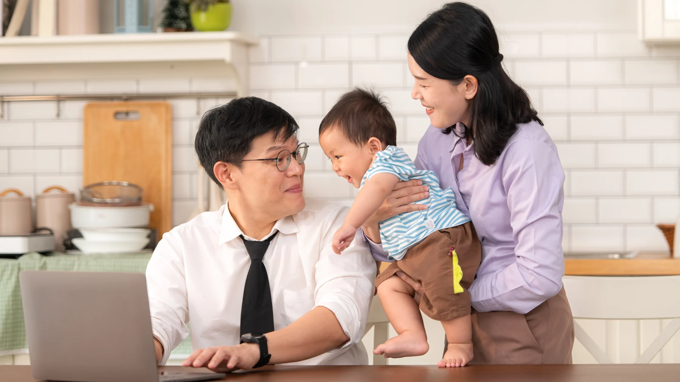 Dad works on a laptop at the kitchen counter while mom lifts their baby toward him for a playful moment.