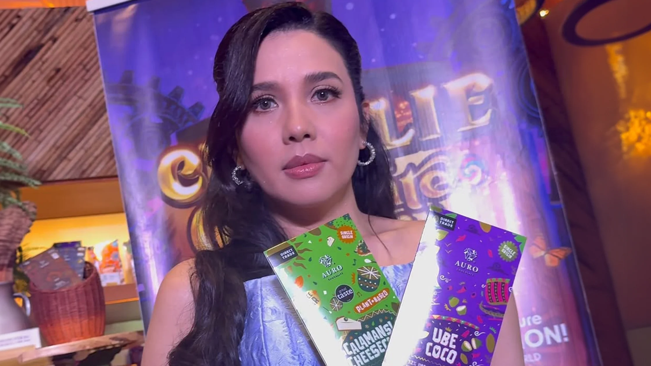 Portrait of a woman with dark hair and hoop earrings, holding bright, colorful product brochures at a booth.