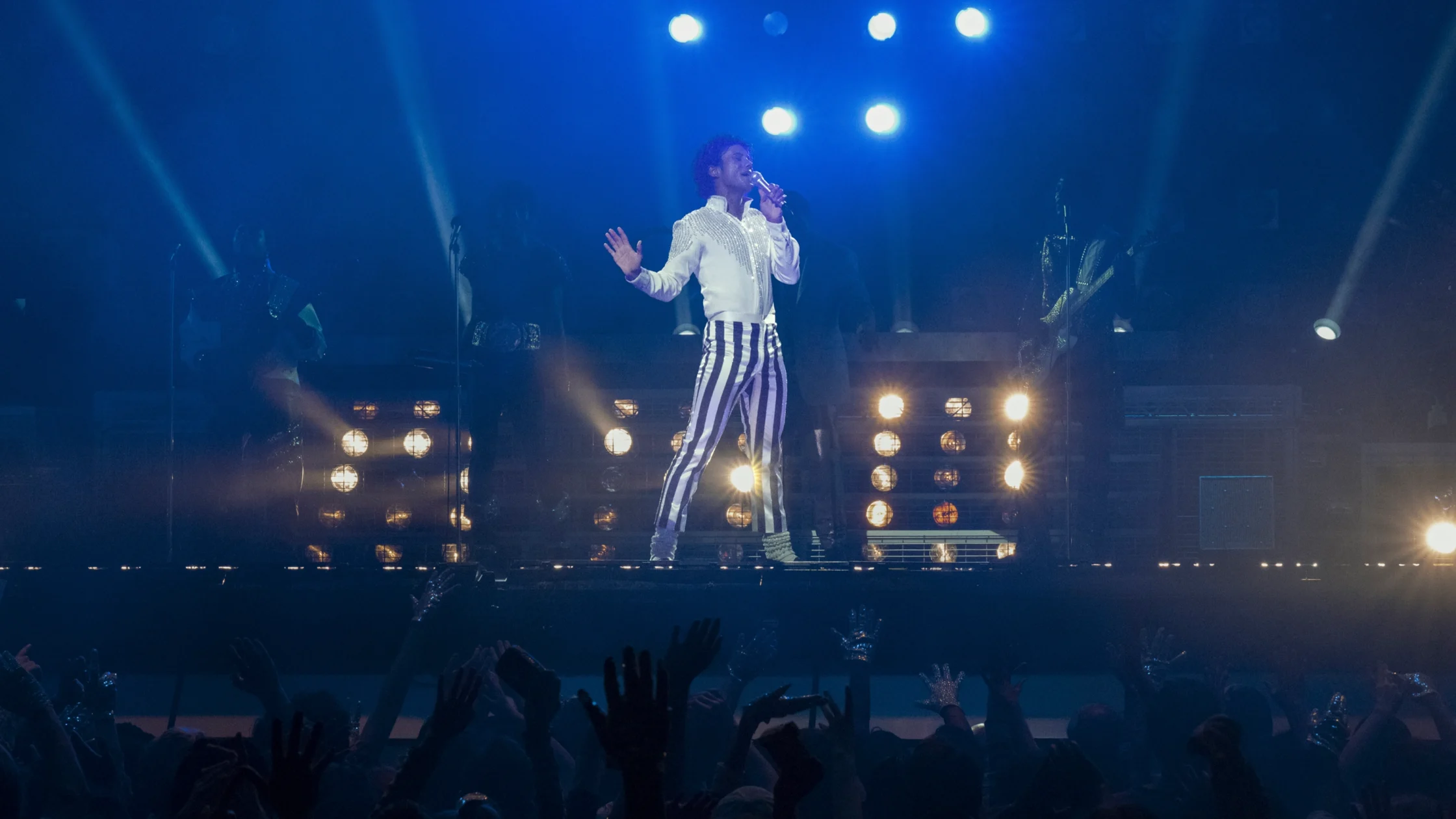 Male singer on a lit stage wearing a white jacket and striped pants, singing into a microphone as blue spotlights shine down and a cheering crowd raises their hands.