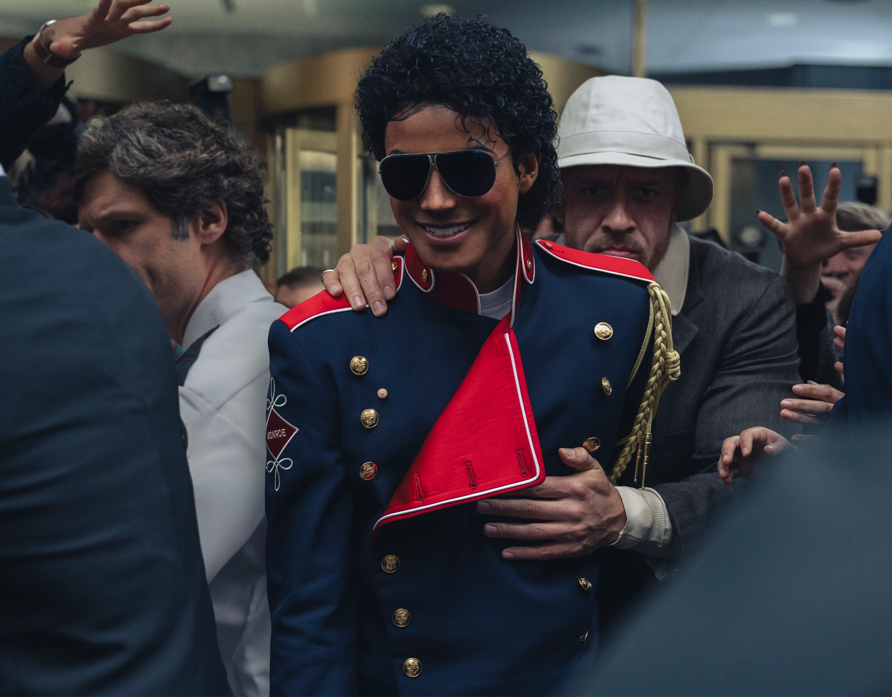 Elvis-inspired performer in a dark blue military-style coat with red sash, wearing sunglasses, being guided by the crowd.