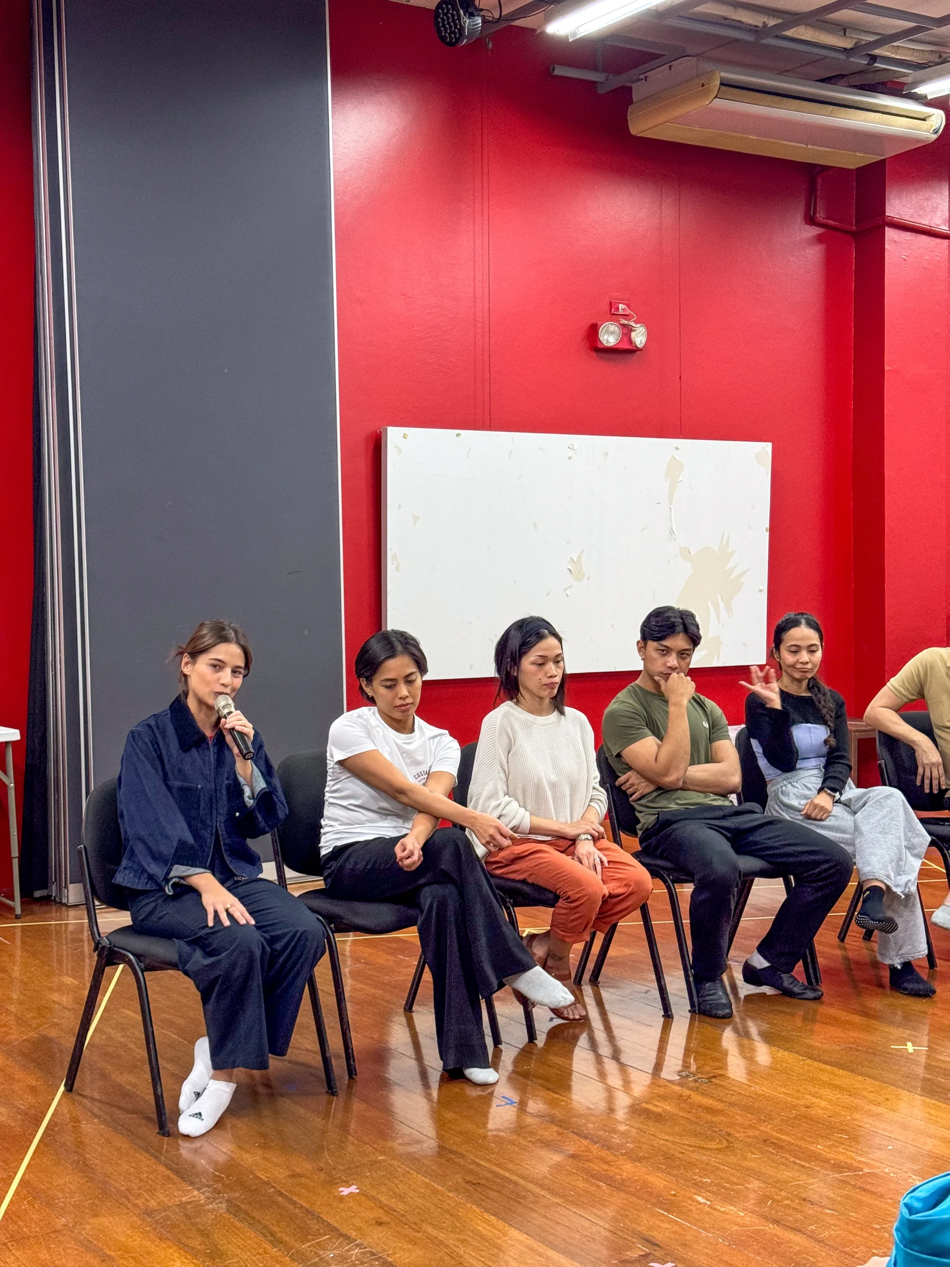 Six people seated in a row in a red-walled studio; the woman on the far left speaks into a microphone, as if moderating a panel.