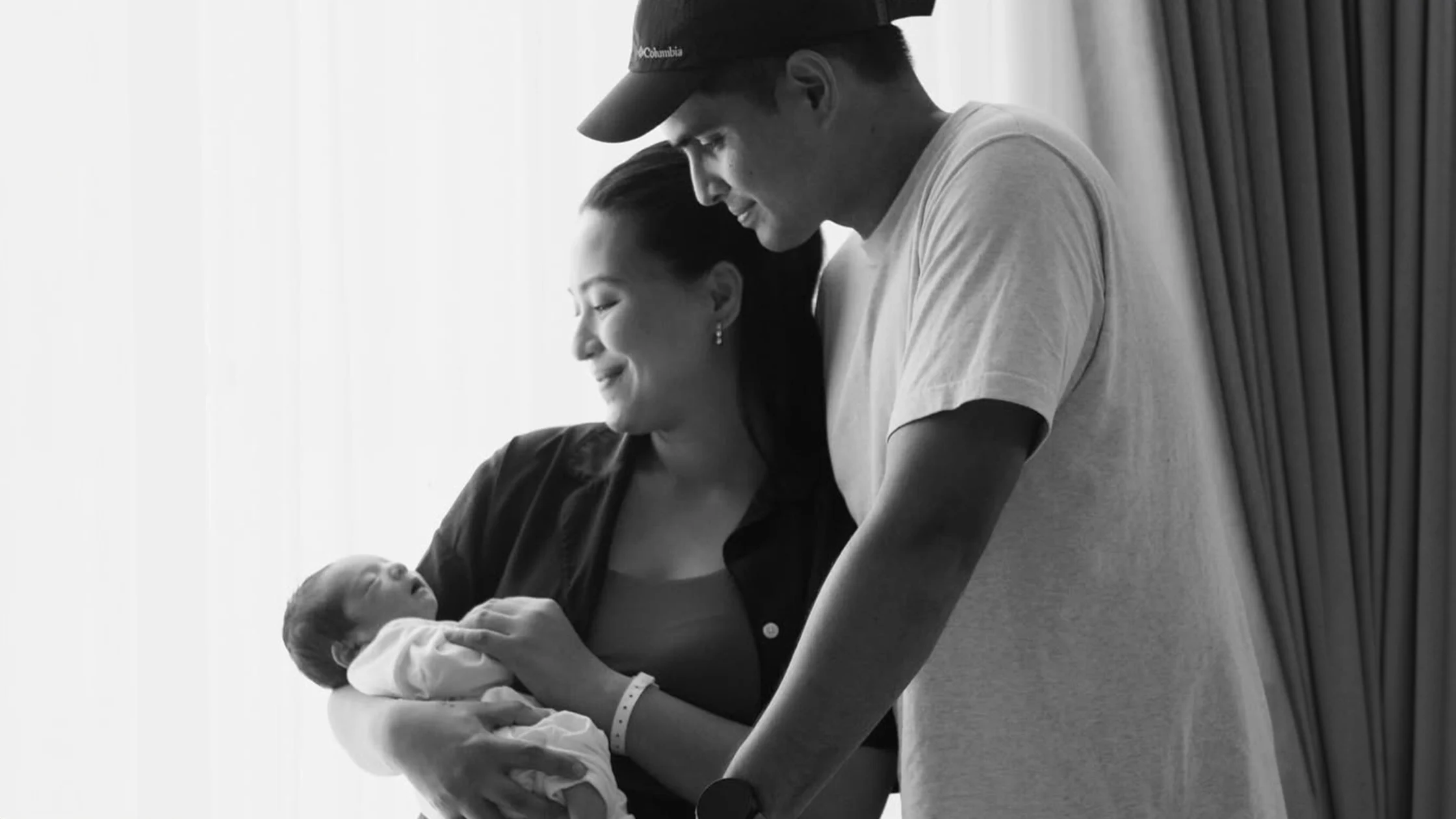 Mother cradles newborn while father stands nearby, smiling in a bright room.