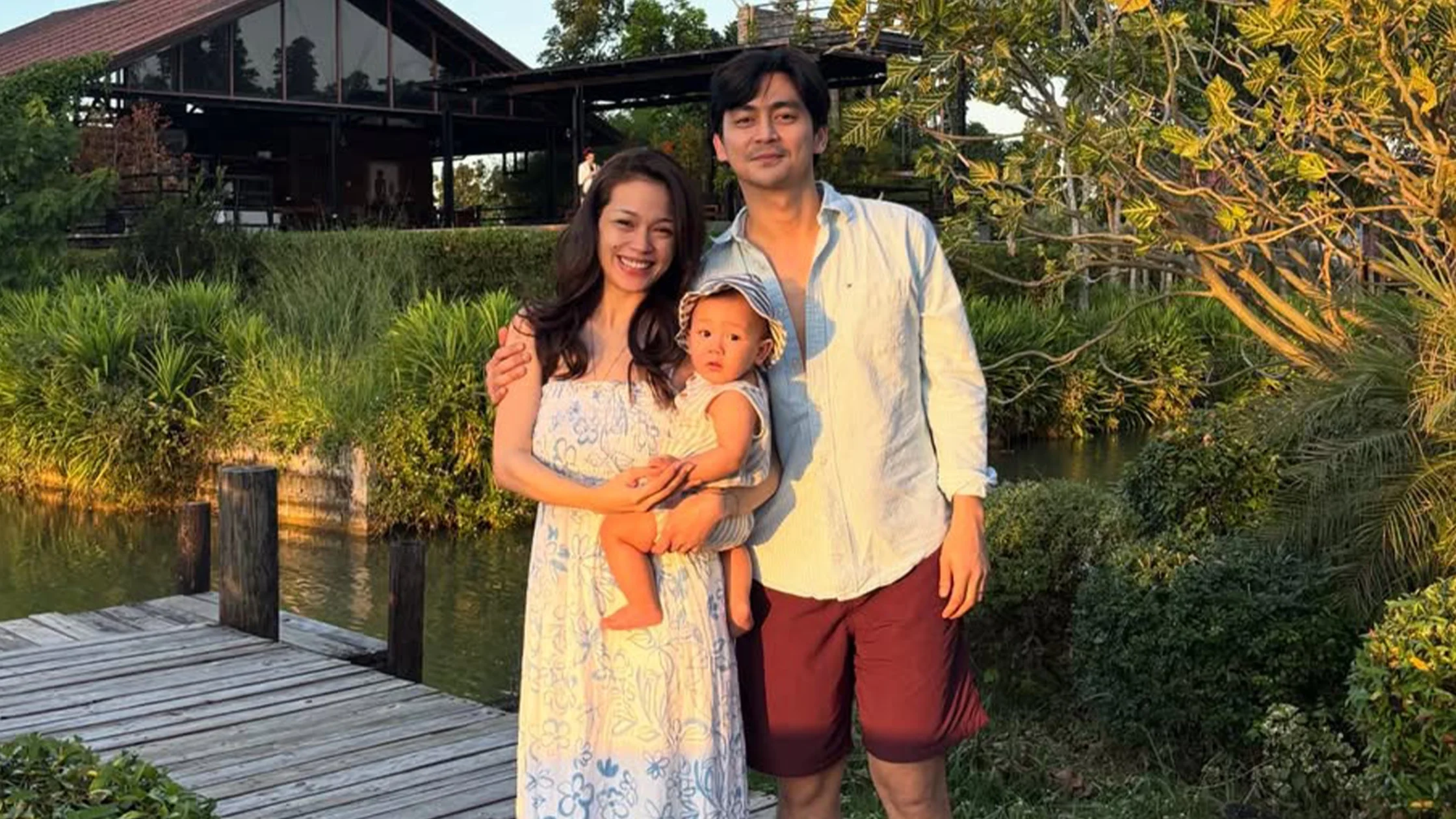 Smiling family of three standing on a wooden dock beside a canal, with a house and lush greenery in the background.