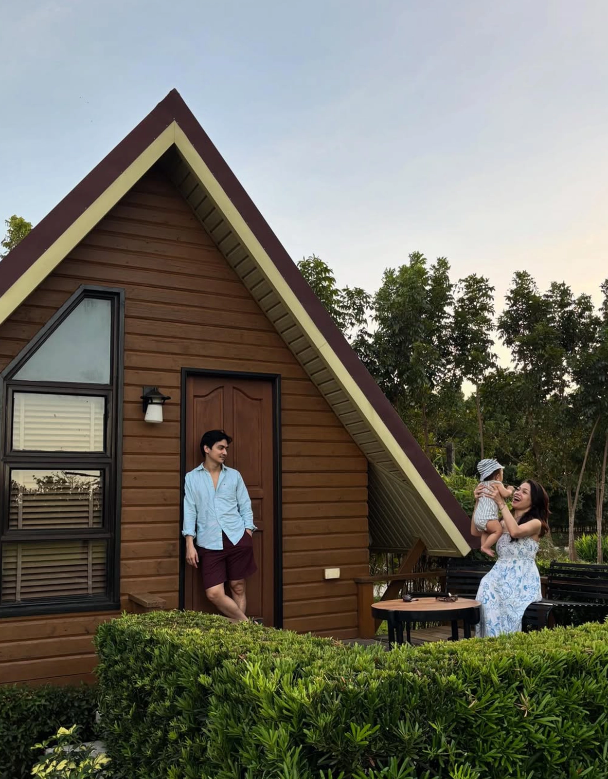 A man wearing a light blue shirt and maroon shorts stands by the wooden A-frame cottage door while a woman in a floral dress lifts a baby outdoors on a patio.