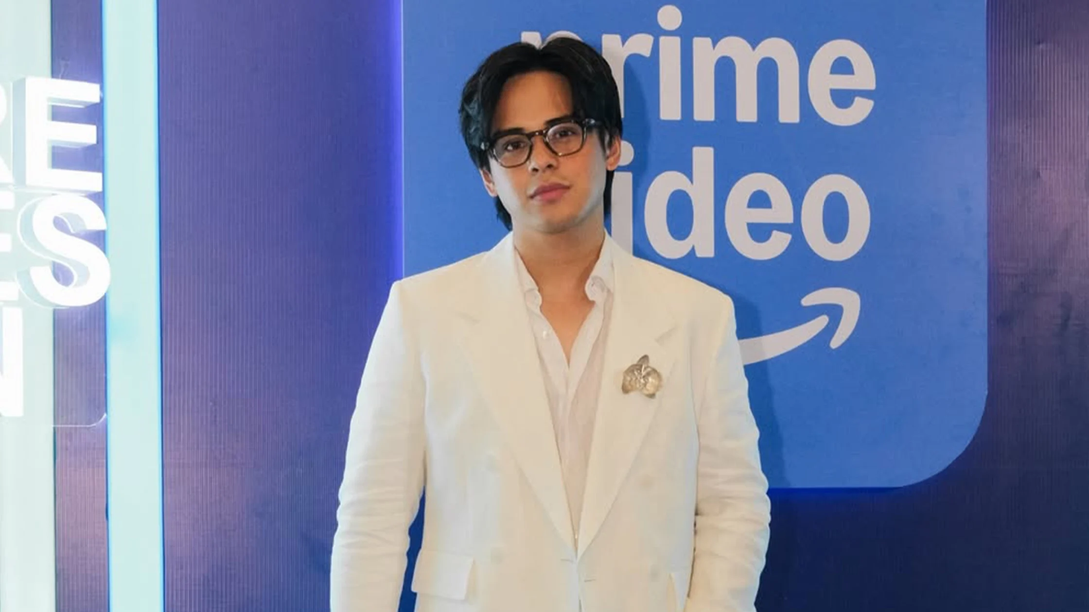 Young man in a white suit and glasses posing in front of a blue Prime Video backdrop.