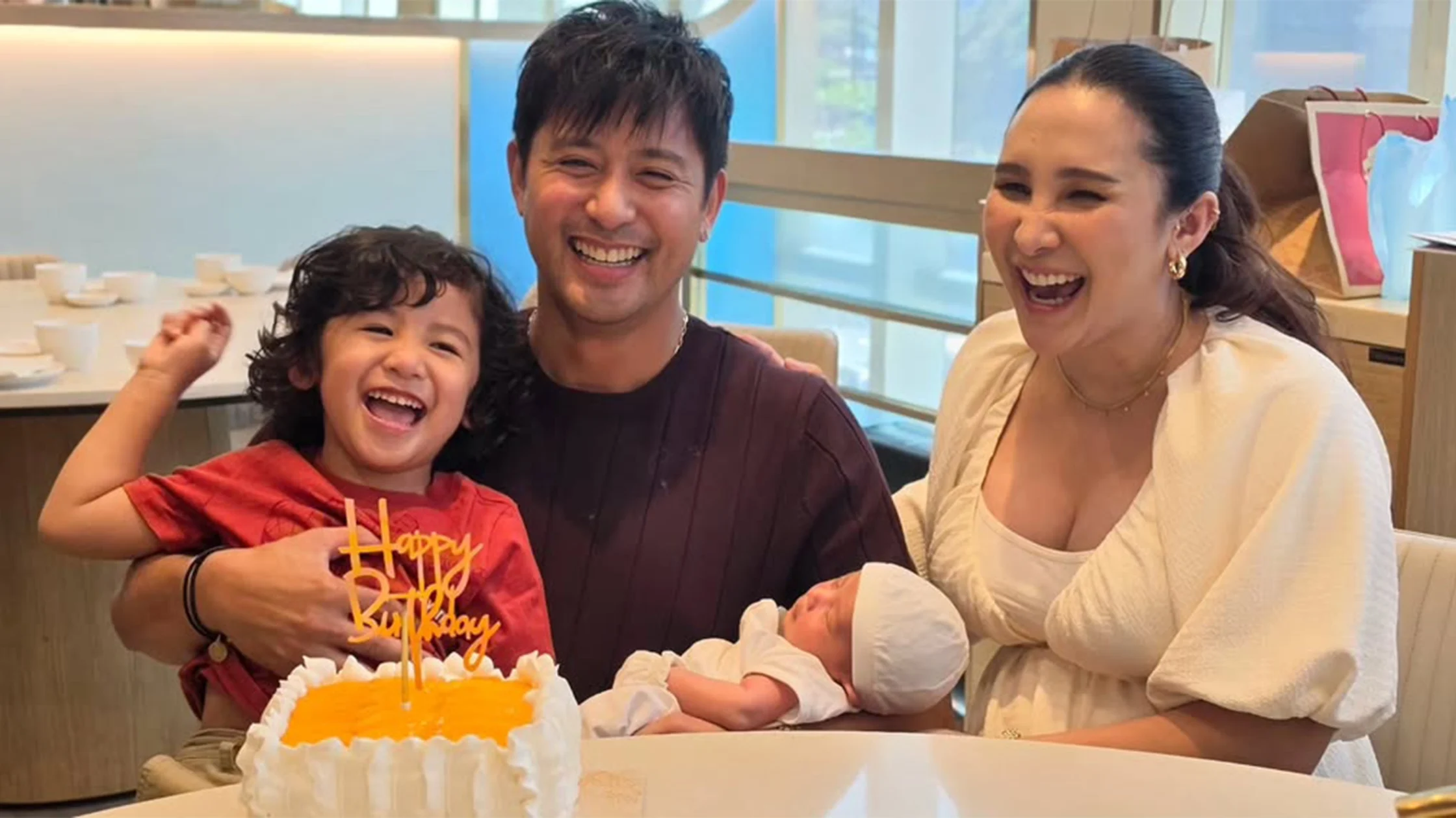 Family birthday scene: a man holds a laughing toddler, a woman smiles with a newborn baby, and a yellow-trost cake sits on the table in front of them.