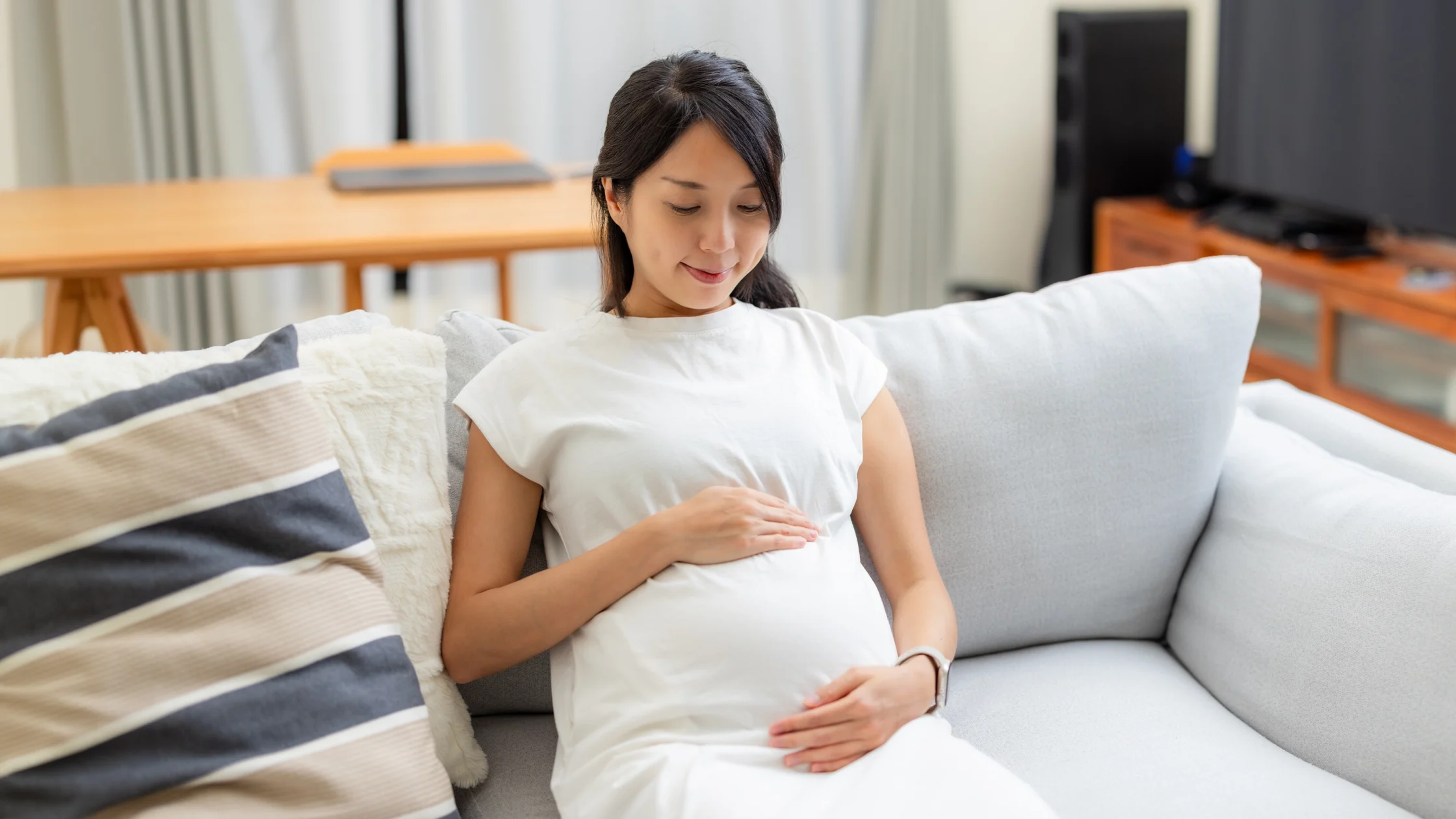 Pregnant woman sits on a light gray sofa at home, hands resting on her baby bump.