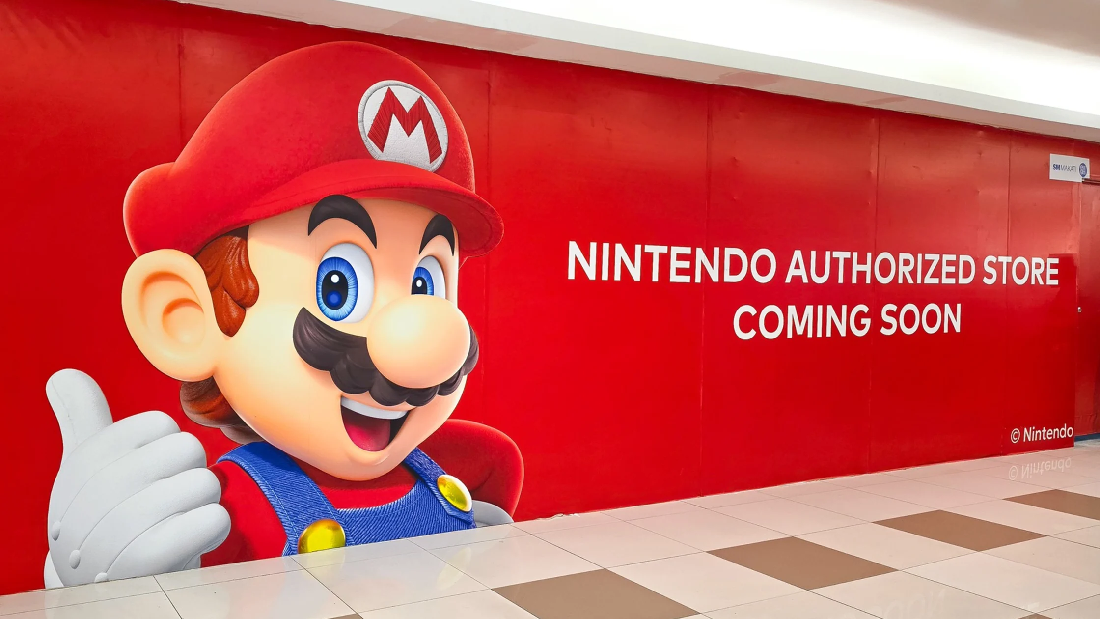 Large Mario character cutout with red hat giving a thumbs-up beside a red wall that reads 'NINTENDO AUTHORIZED STORE COMING SOON'.