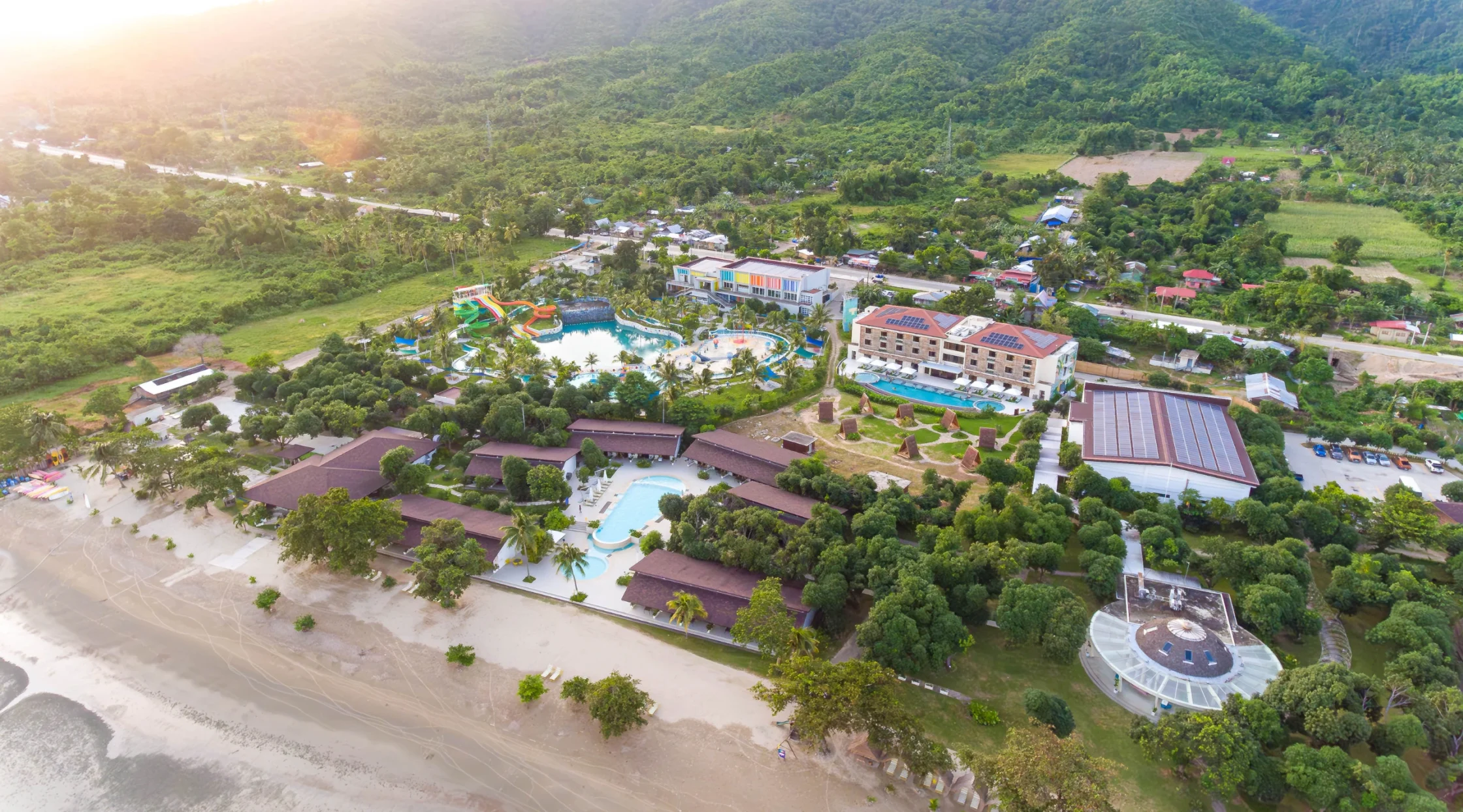 Aerial view of a tropical resort with a large pool, colorful water slides, palm trees, and surrounding buildings by the beach.