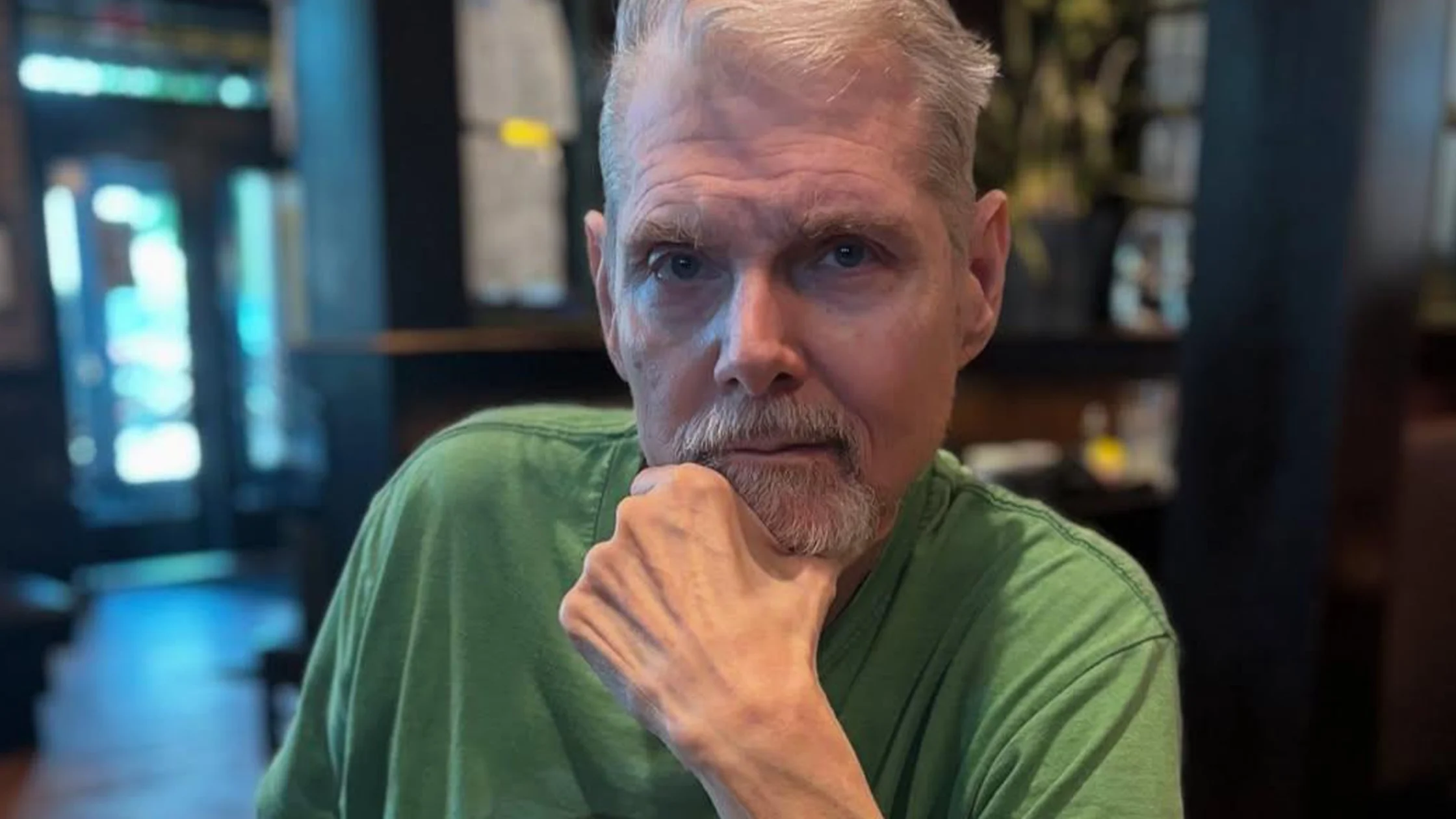 Close-up of a man with gray hair and beard wearing a green shirt, resting his chin on his hand in a cafe setting.