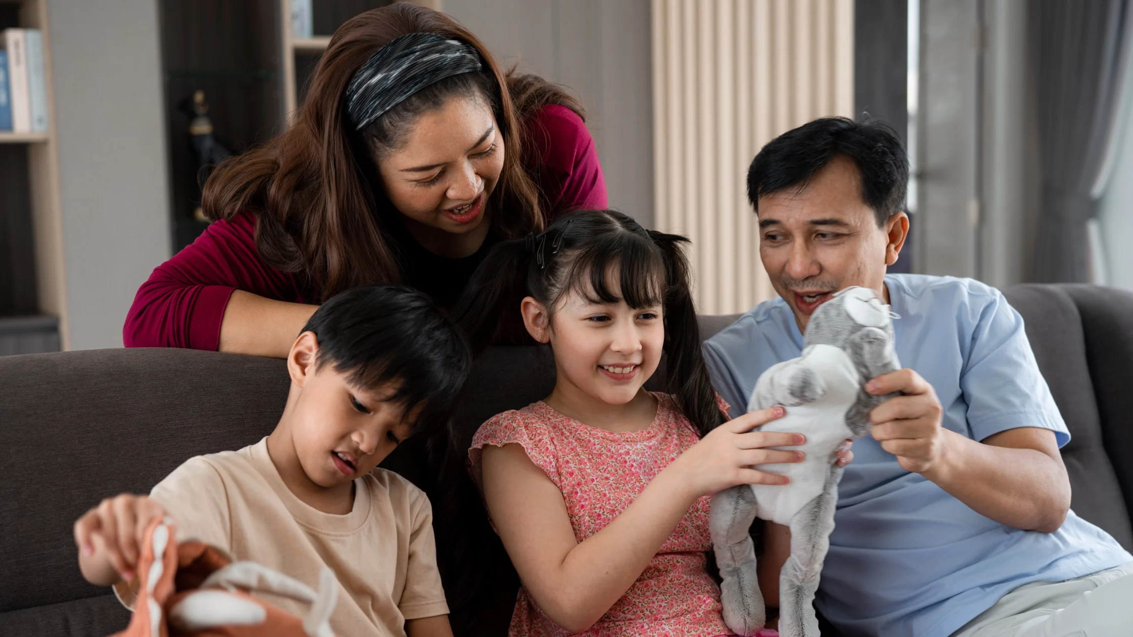 A family of four sits on a sofa, smiling as they play with a gray stuffed toy together indoors.