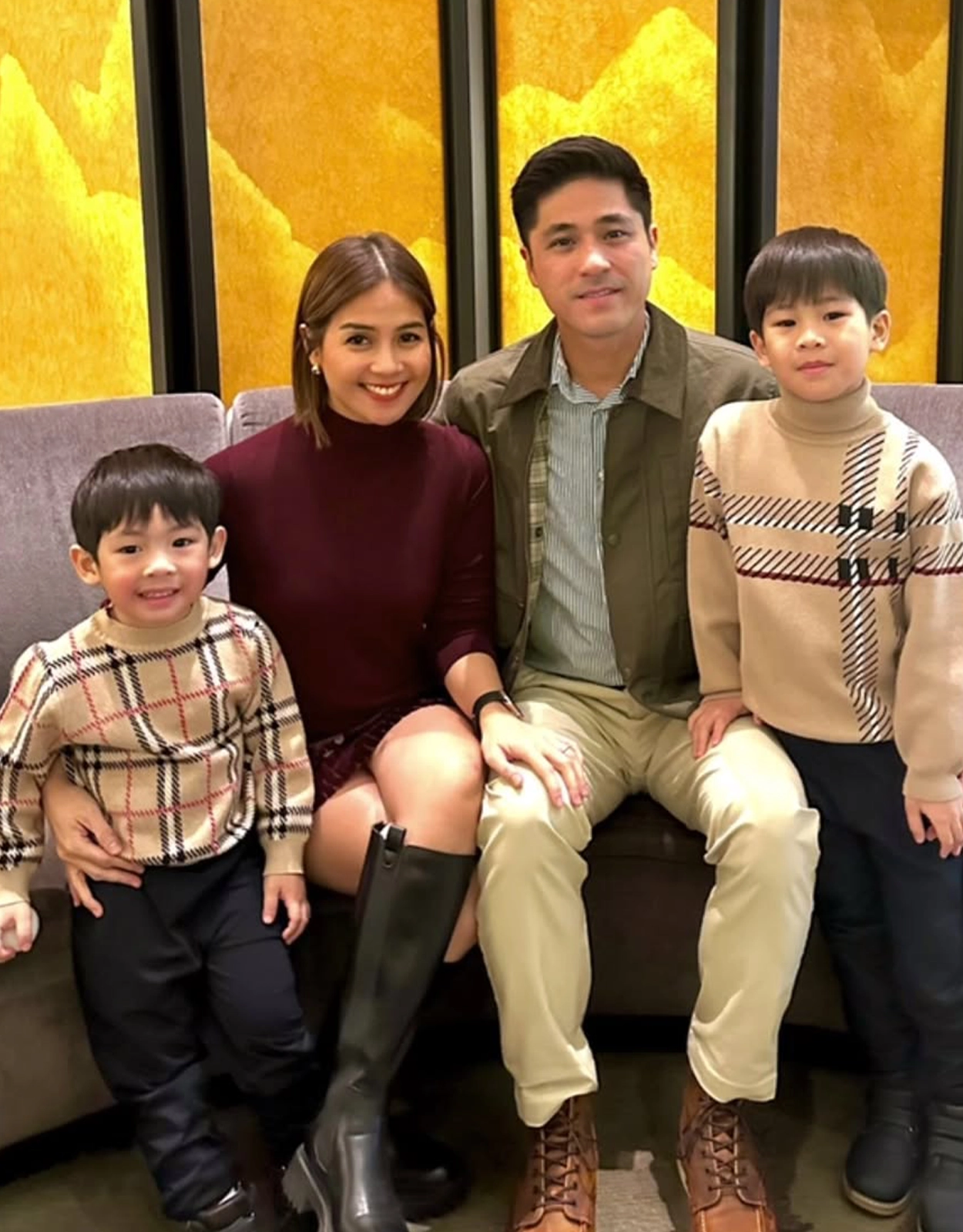 Family of four posing on a sofa indoors, mom in burgundy dress, dad in a green jacket, two boys in matching plaid sweaters.