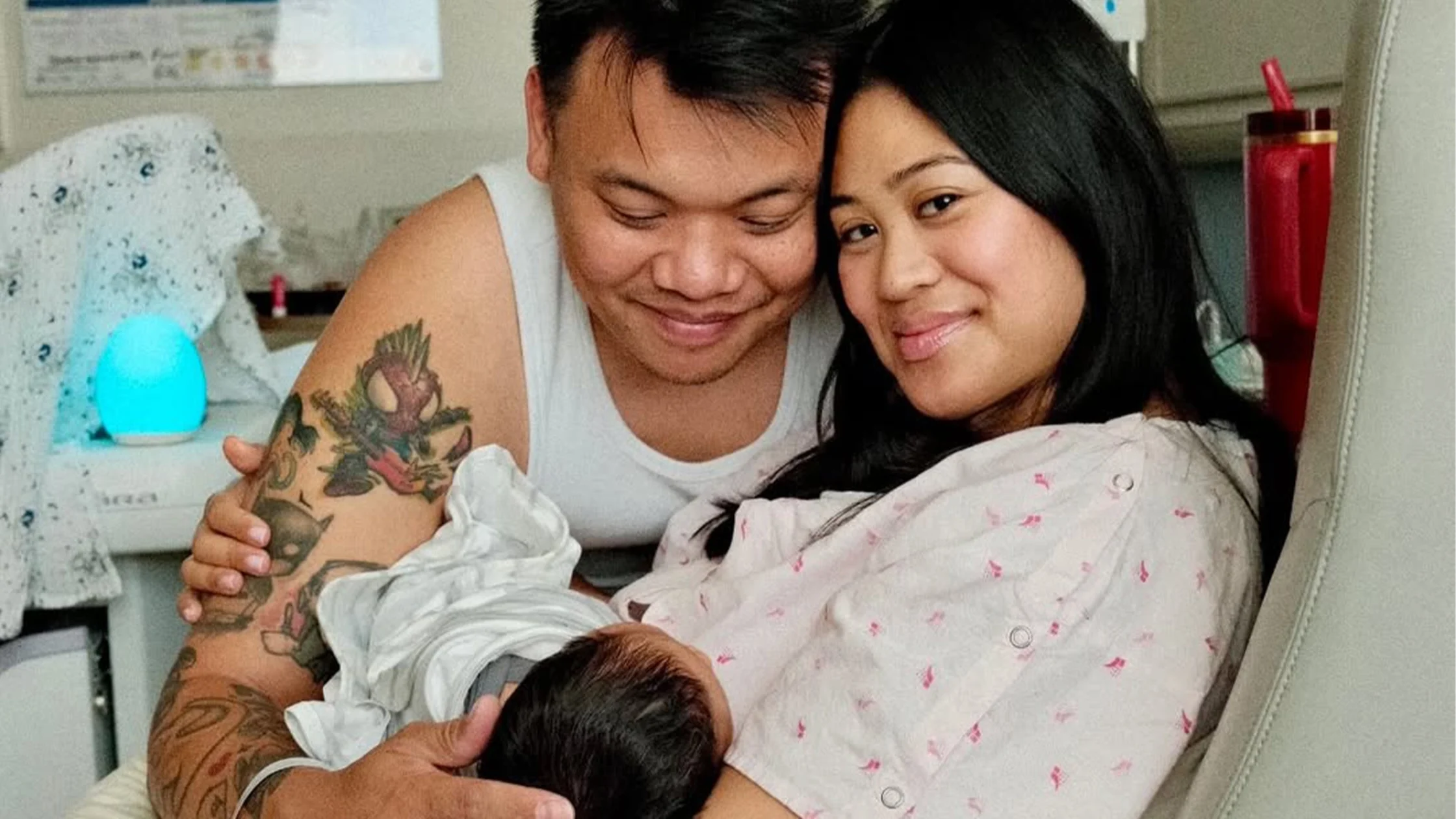 Parents in a hospital room smiling as they cradle a newborn wrapped in a blanket in their arms (father with tattoos).