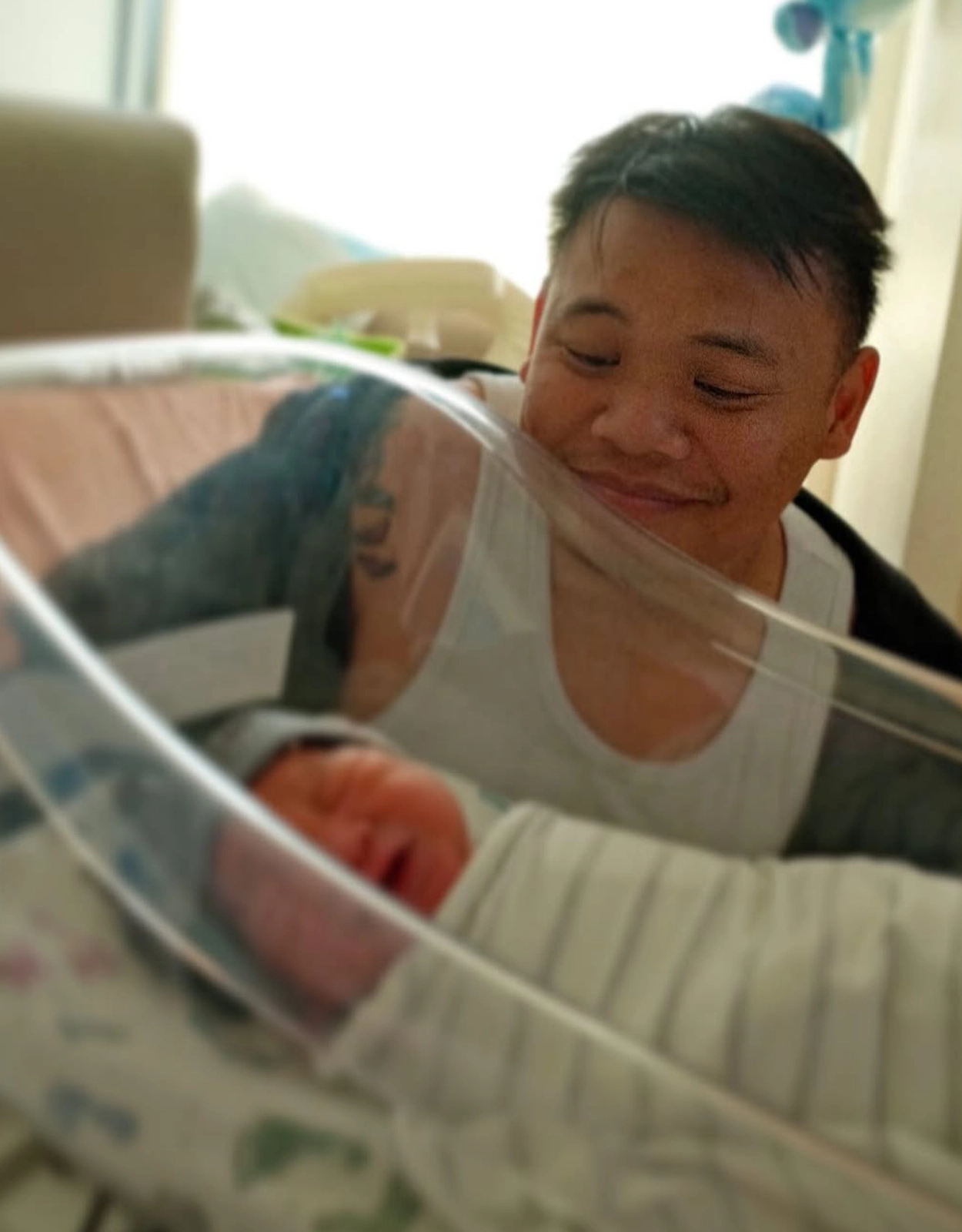 Father smiling at newborn baby in a hospital bassinet, baby wrapped in a blanket on a pale bed.