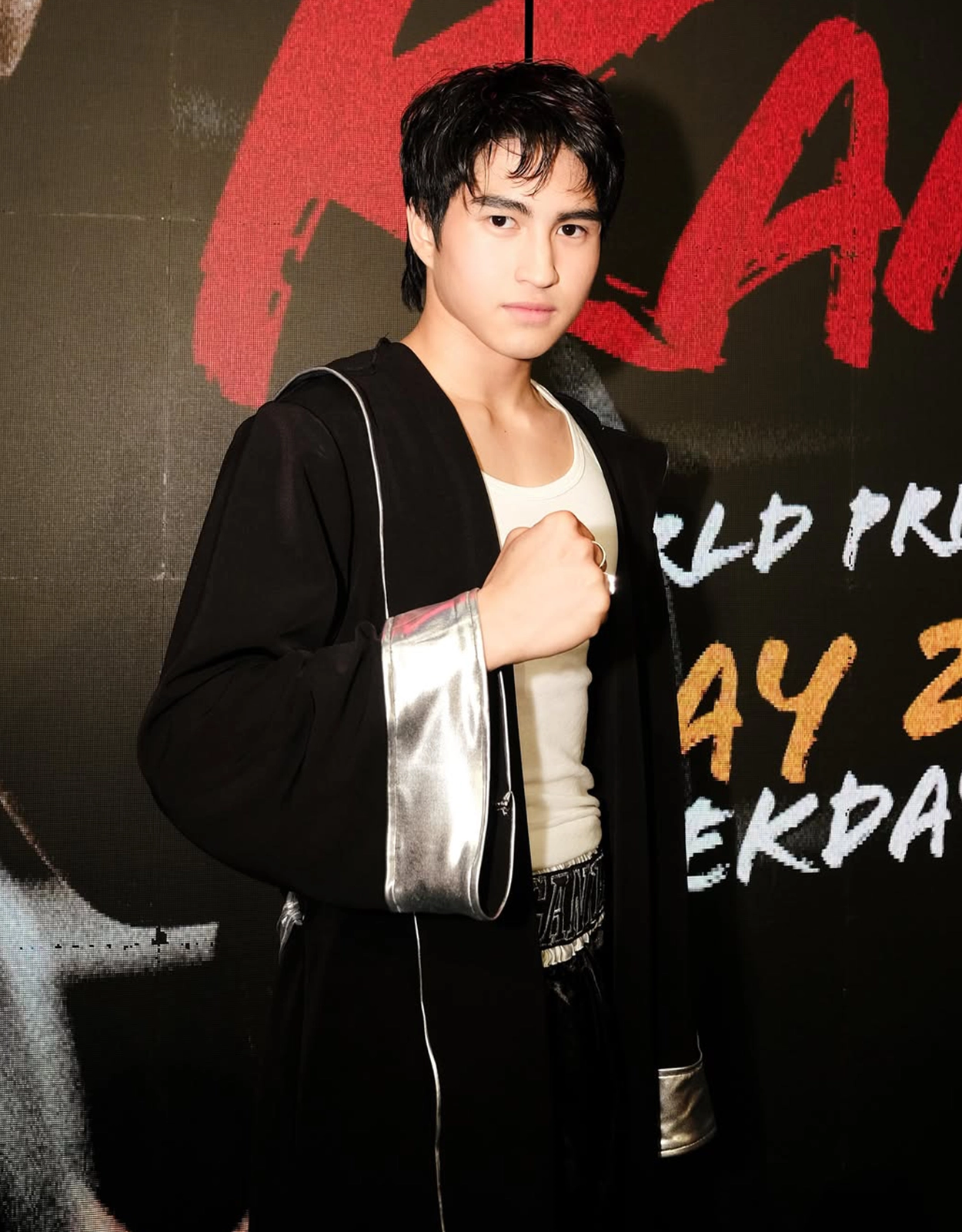 Young man in a black robe with silver trim, posing with a raised fist at a red-carpet backdrop.