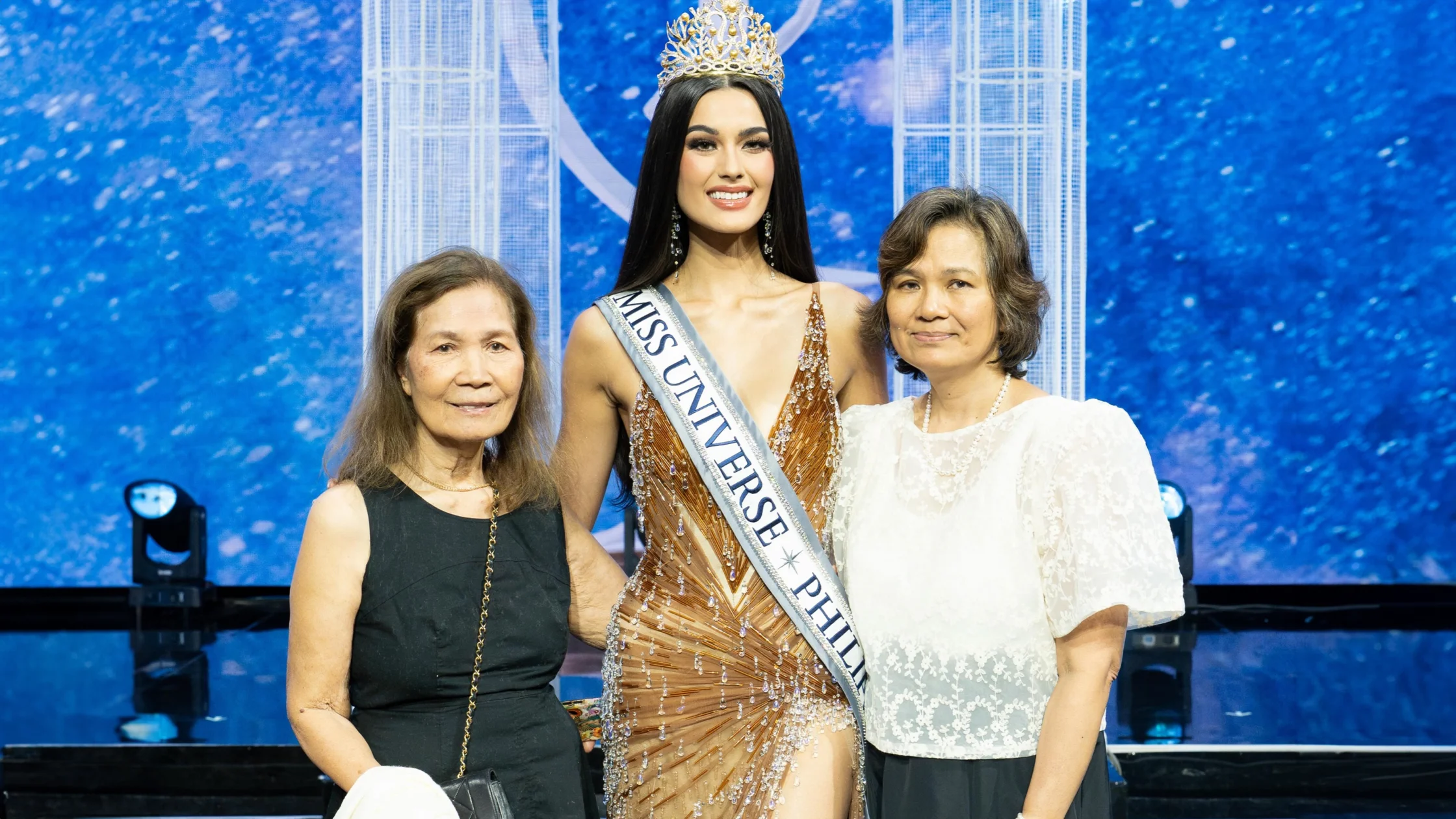 Miss Universe winner in a jeweled orange gown and crown, flanked by two smiling women on a stage with a blue backdrop and sash reading 'MISS UNIVERSE'.