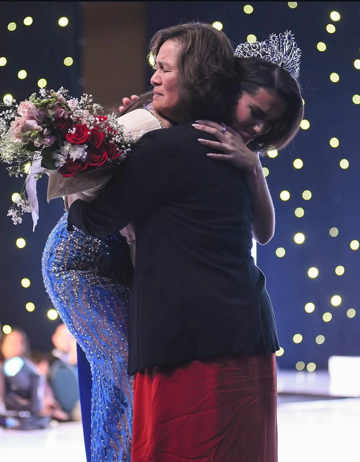 Two people hug on stage; one wearing a sparkling gown and a crown, holding a bouquet of pink and red roses as lights glow behind them.