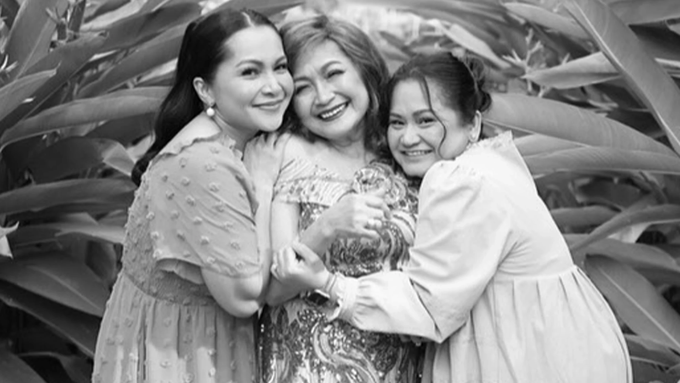Three women in dresses hug and smile together outdoors among large tropical leaves.