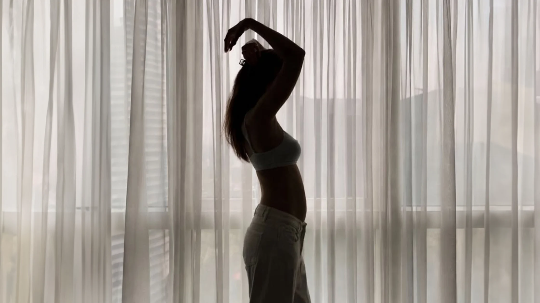 Silhouette of a person in a sports bra and jeans stretching with arms raised overhead in front of sheer curtains.