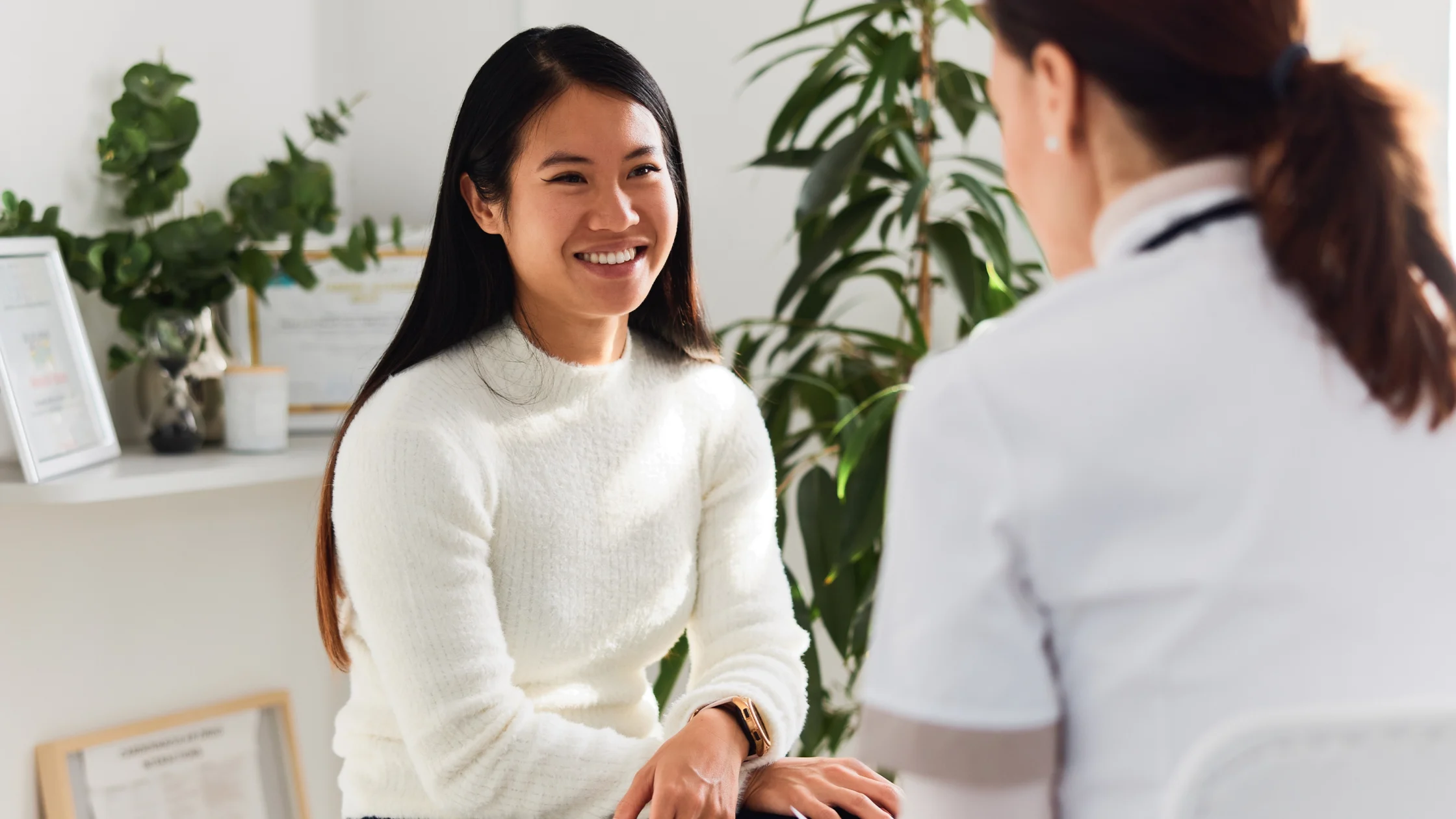 Smiling woman in a white sweater sits facing a coworker in a white coat in a bright office setting with plants in the background.