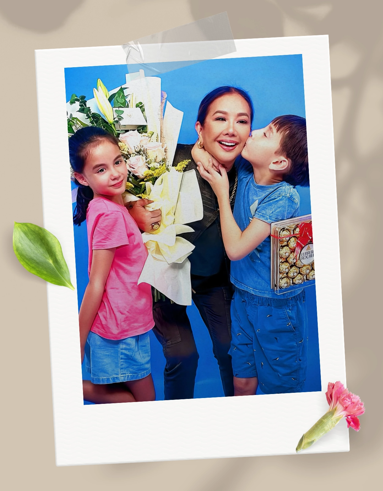 Smiling woman posing with two children, who kiss her cheek, while she holds a large bouquet of flowers against a blue background.