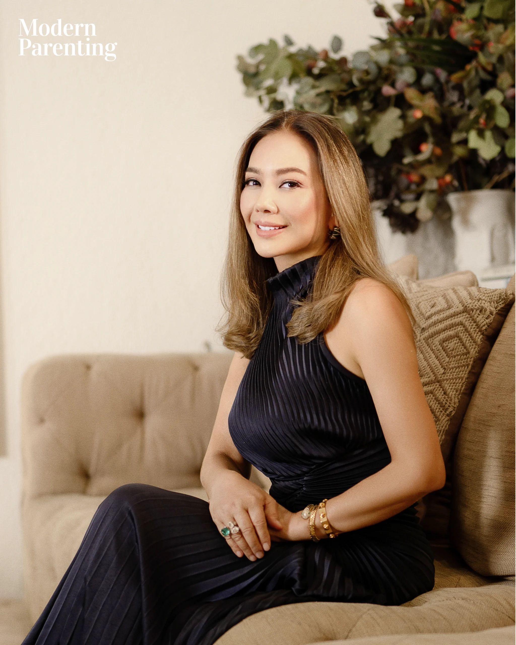 Smiling woman in a dark pleated dress sits on a beige sofa, with a leafy plant behind.