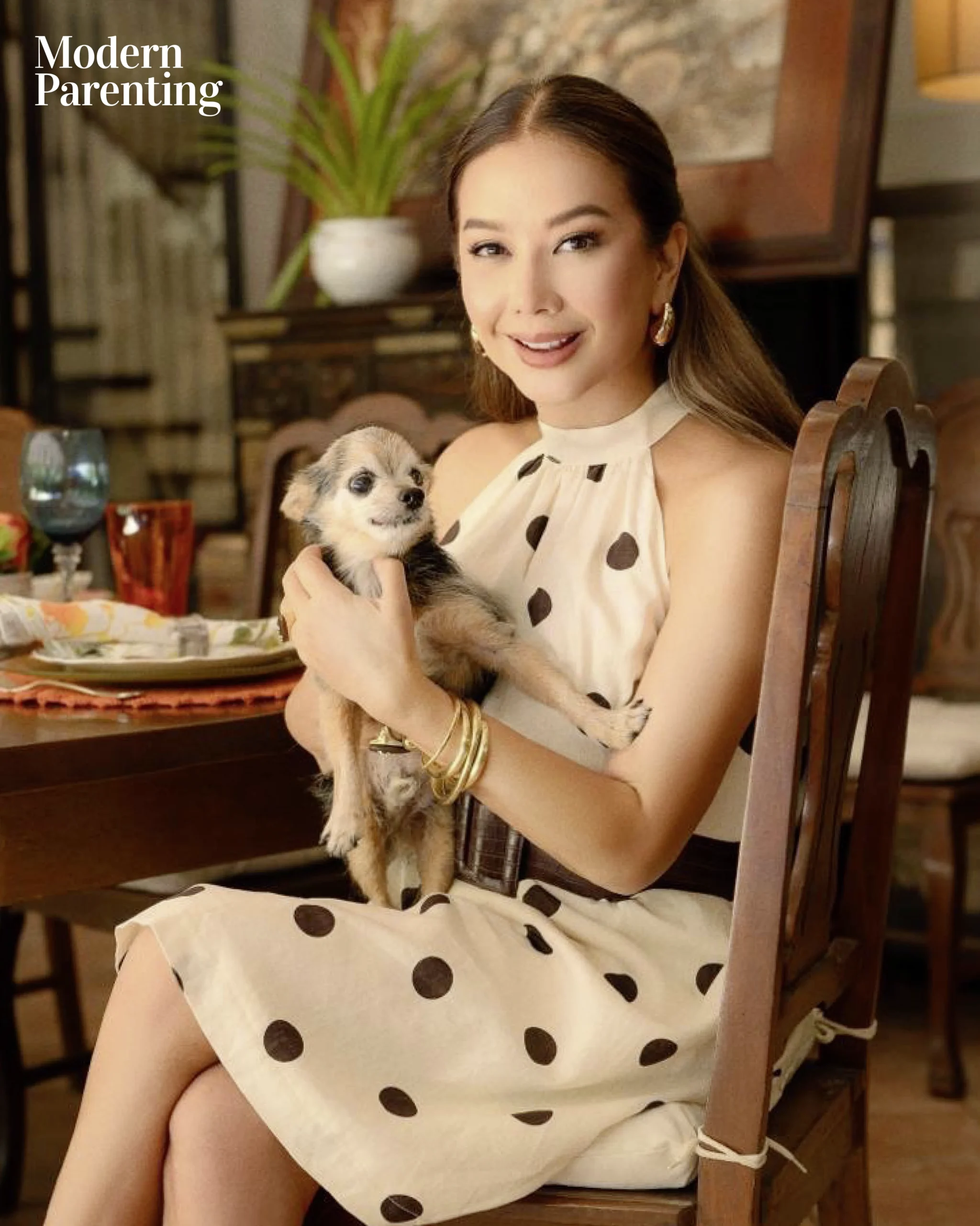 Woman in a cream polka-dot dress sits at a dining table, holding a small fluffy dog; 'Modern Parenting' logo in the corner of the image.