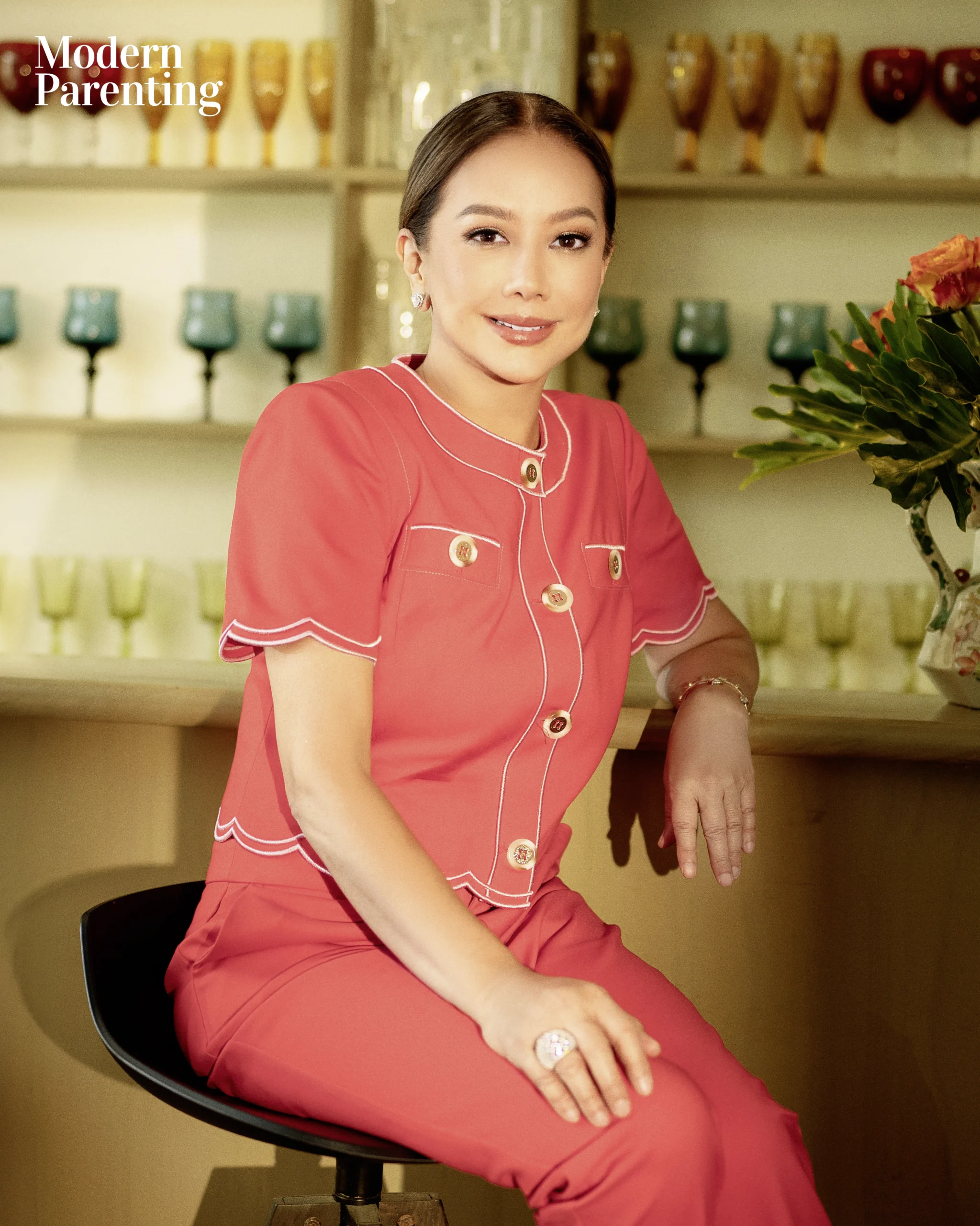 Woman in coral outfit sits at a bar, smiling, with flowers on the counter and glassware in the background.