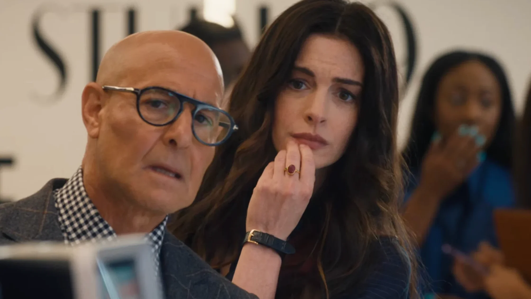 Two professionals at a meeting: an older man with blue-rimmed glasses and a checkered shirt, and a younger woman resting her chin in thought with a blurred colleague in the background.