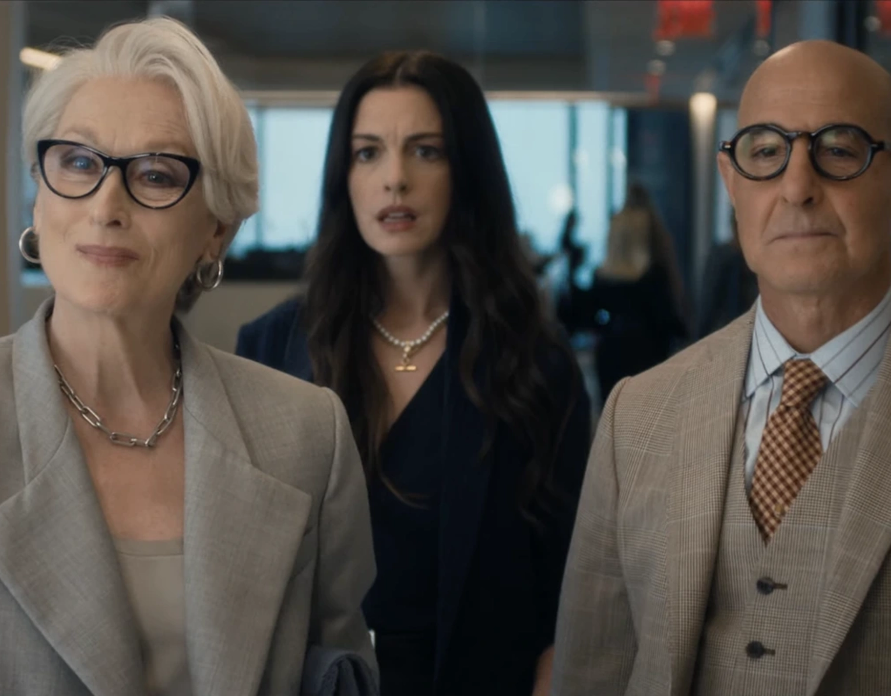 Three business professionals in a modern office, two older adults in beige suits flanking a younger woman in a dark blazer, looking toward the camera in the foreground.