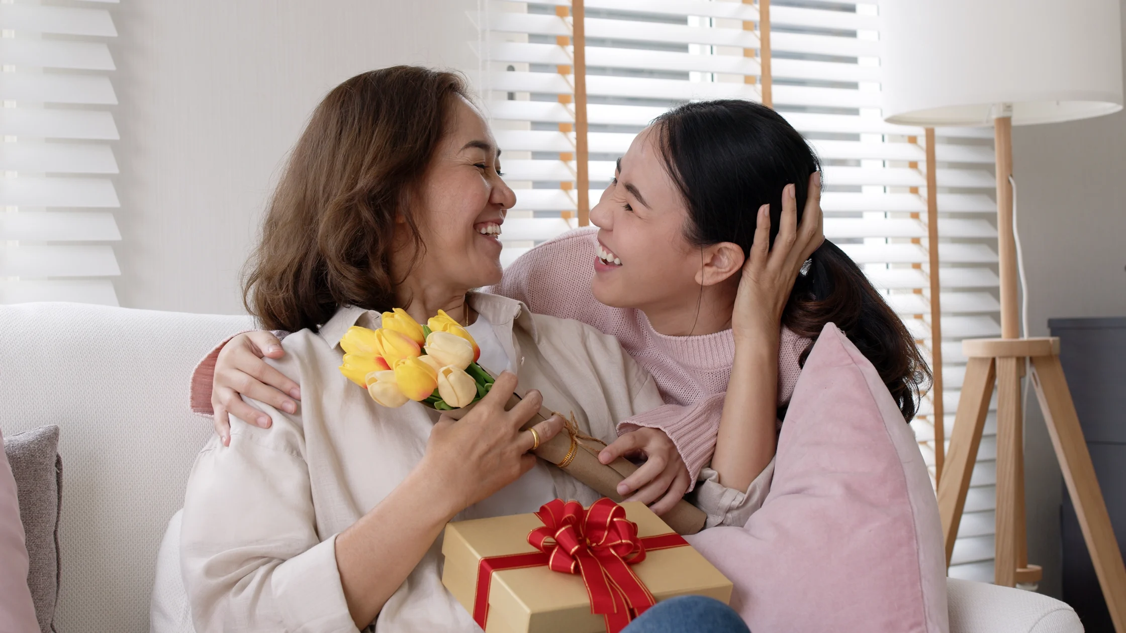 Two women smiling and hugging on a couch, holding a bouquet of yellow tulips and a wrapped gift with a red bow in a bright living room.
