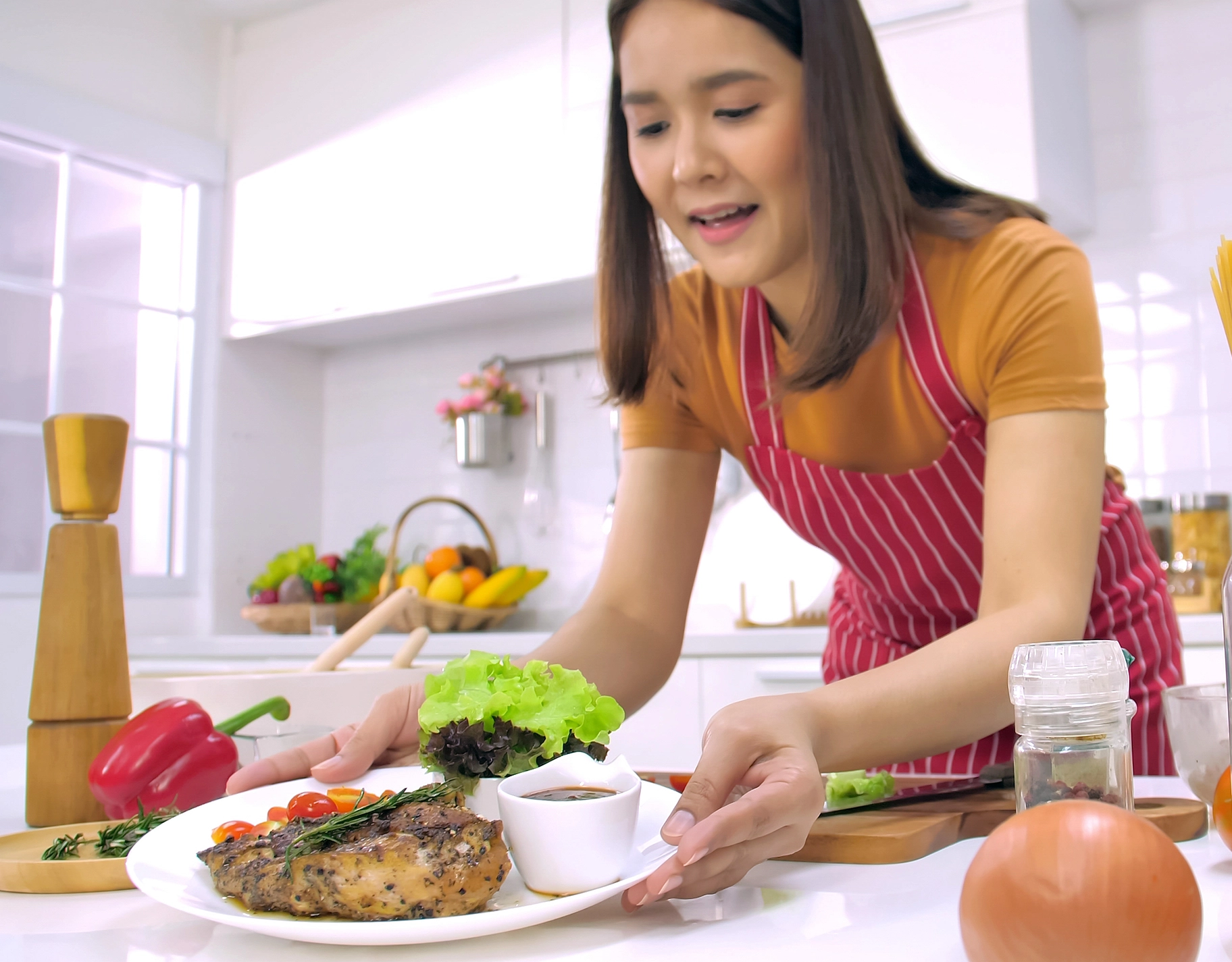 Woman in a red-striped apron garnishes a plate of steak with greens in a bright kitchen.