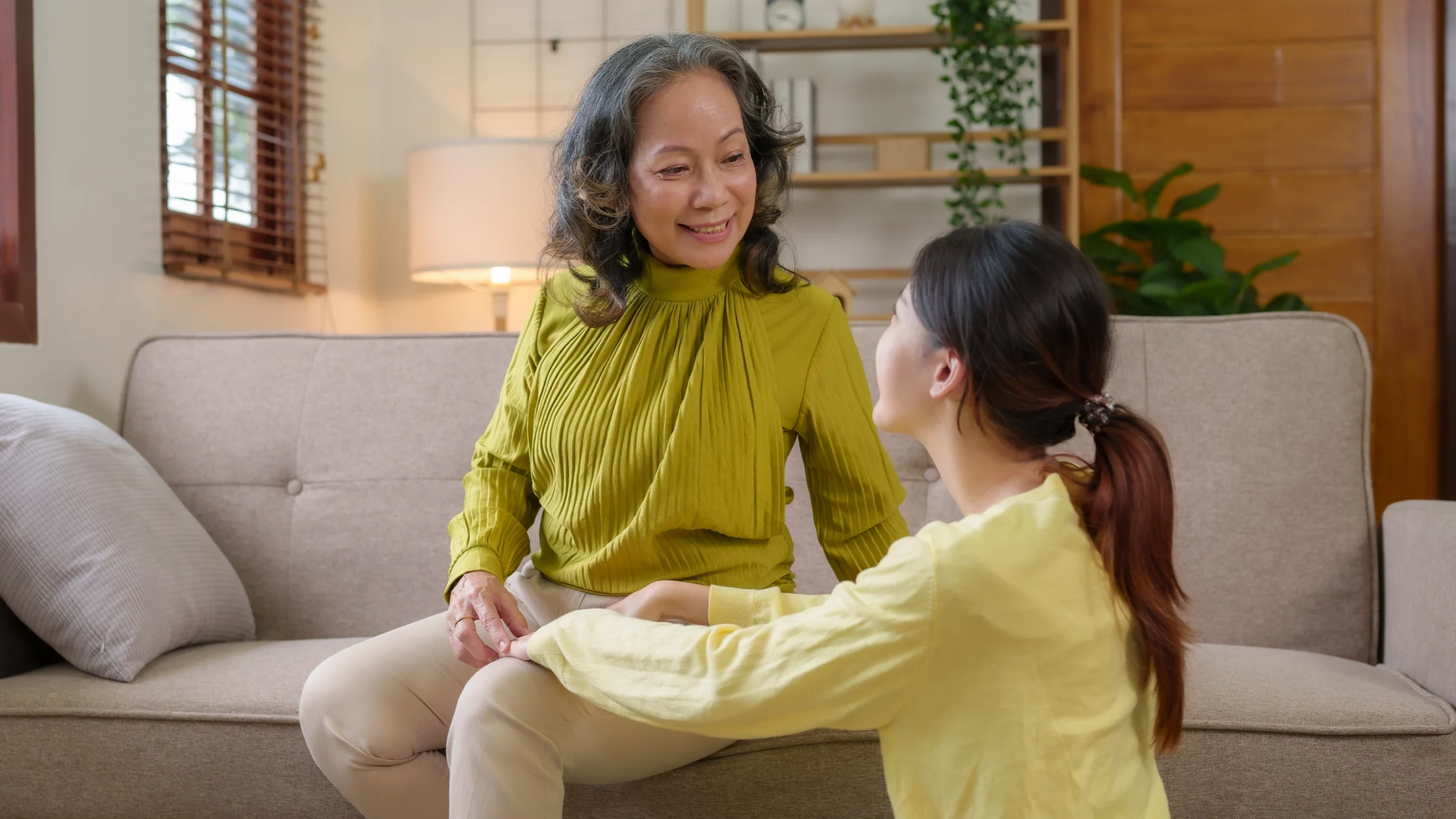 Older woman in a green blouse smiles at a young girl in a yellow top as they sit closely on a beige sofa in a cozy living room.