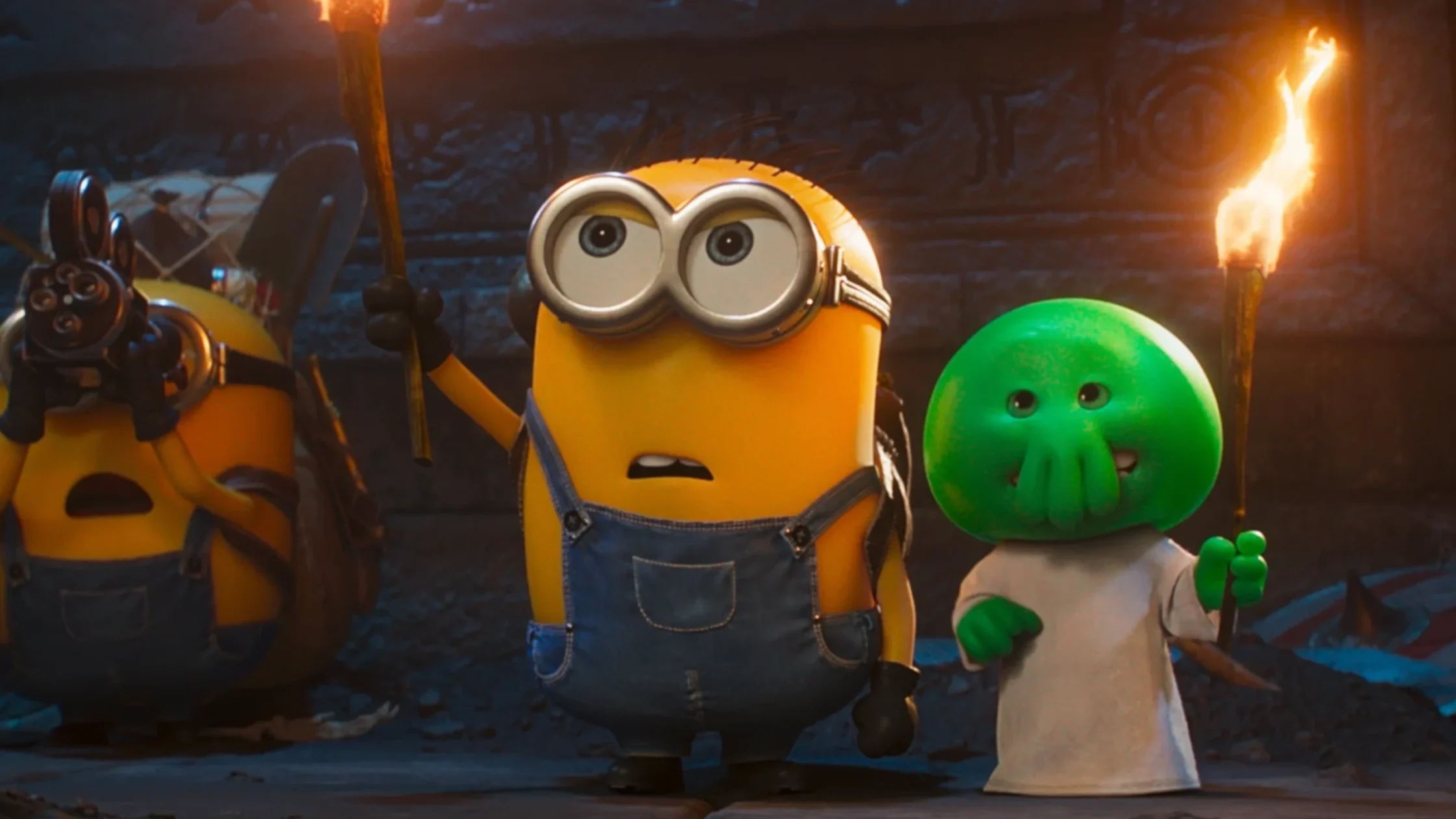 Yellow minion wearing goggles and blue overalls with a green alien, both holding torches in a dark cave. May show a tense or adventurous moment.