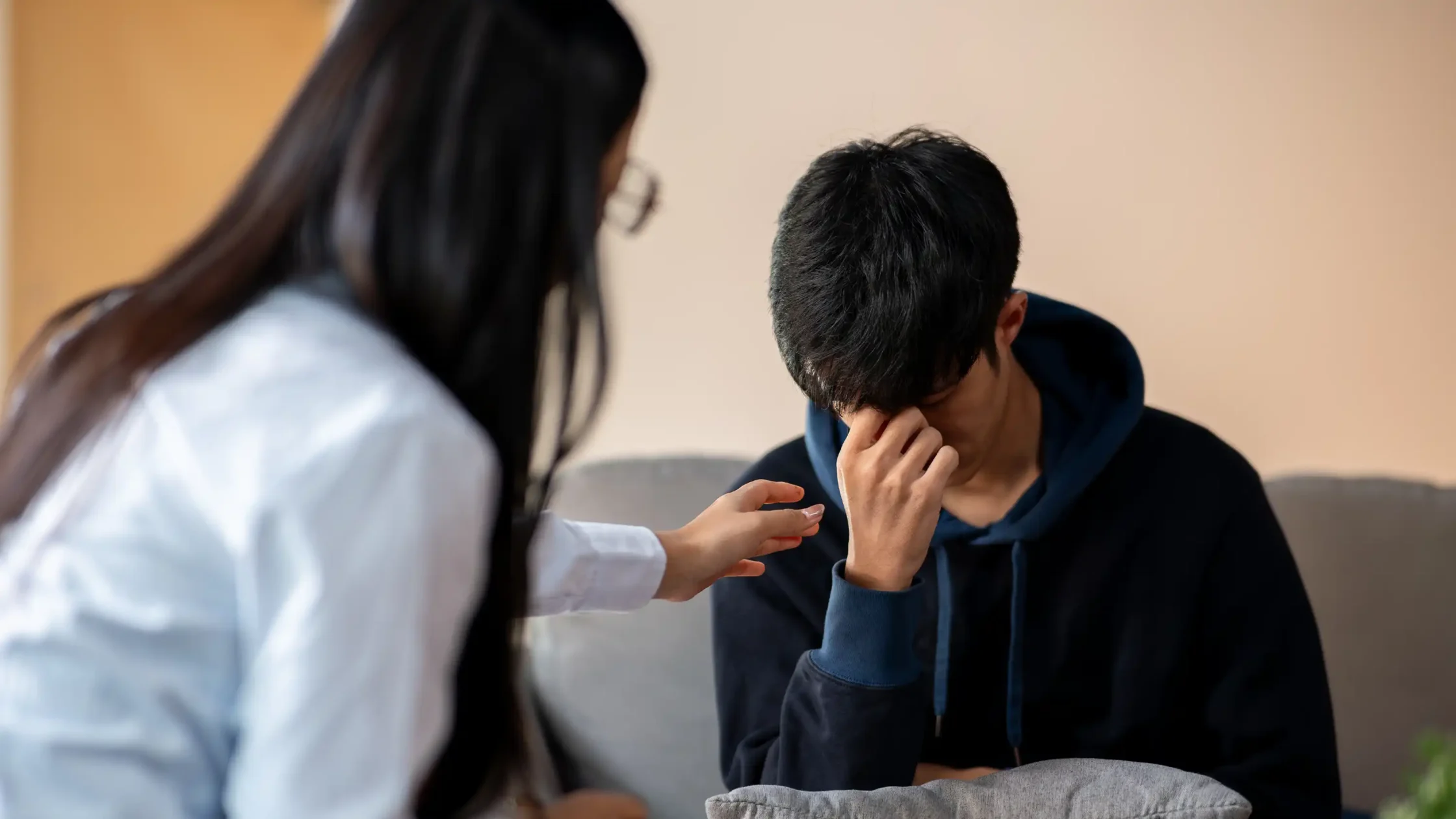 Young man sitting with head down in distress as a woman reaches out to comfort him on a couch.