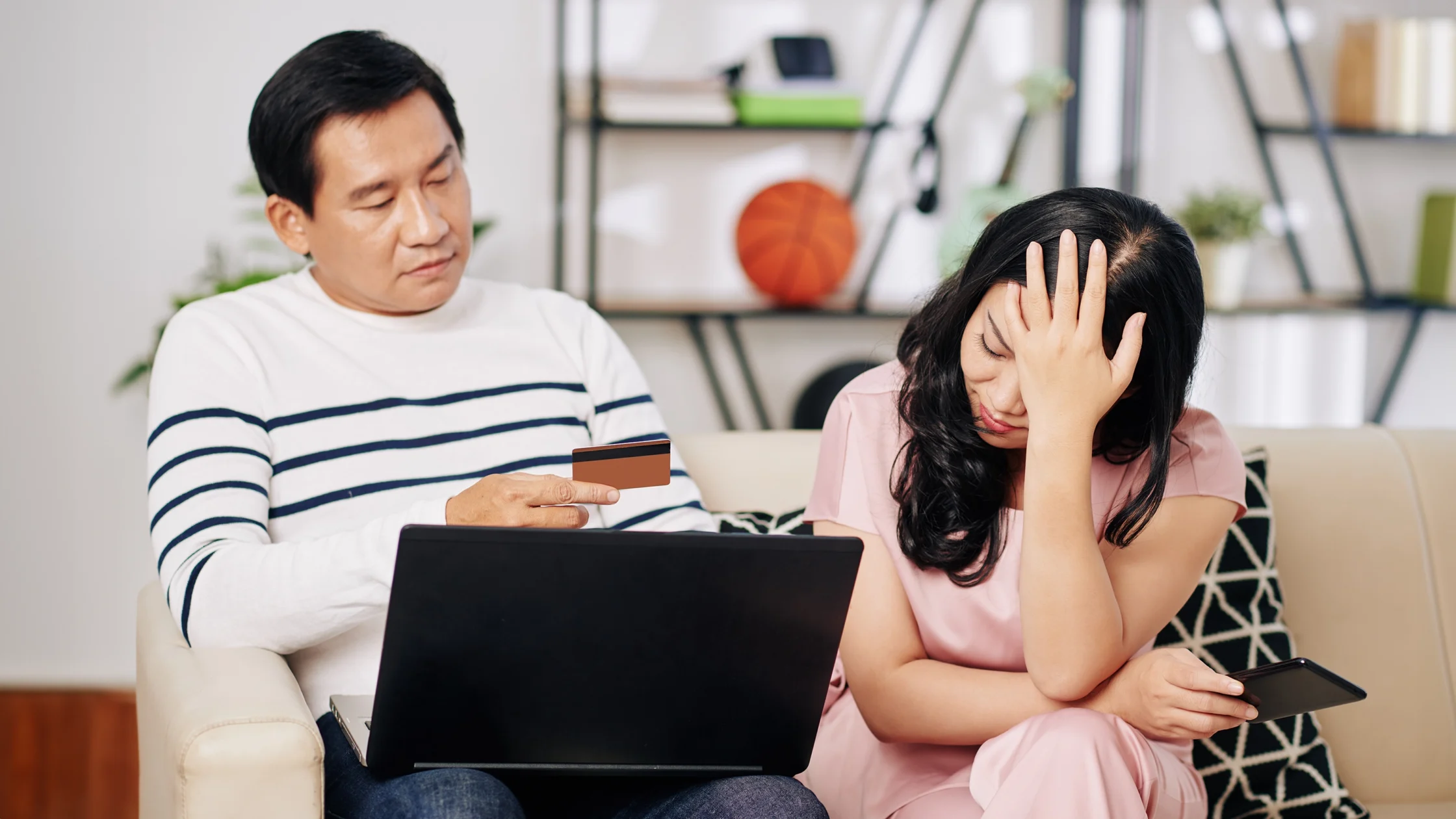 Man wearing a striped shirt shows a credit card to a laptop while a woman in pink sits next to him, her hand on her forehead in distress.