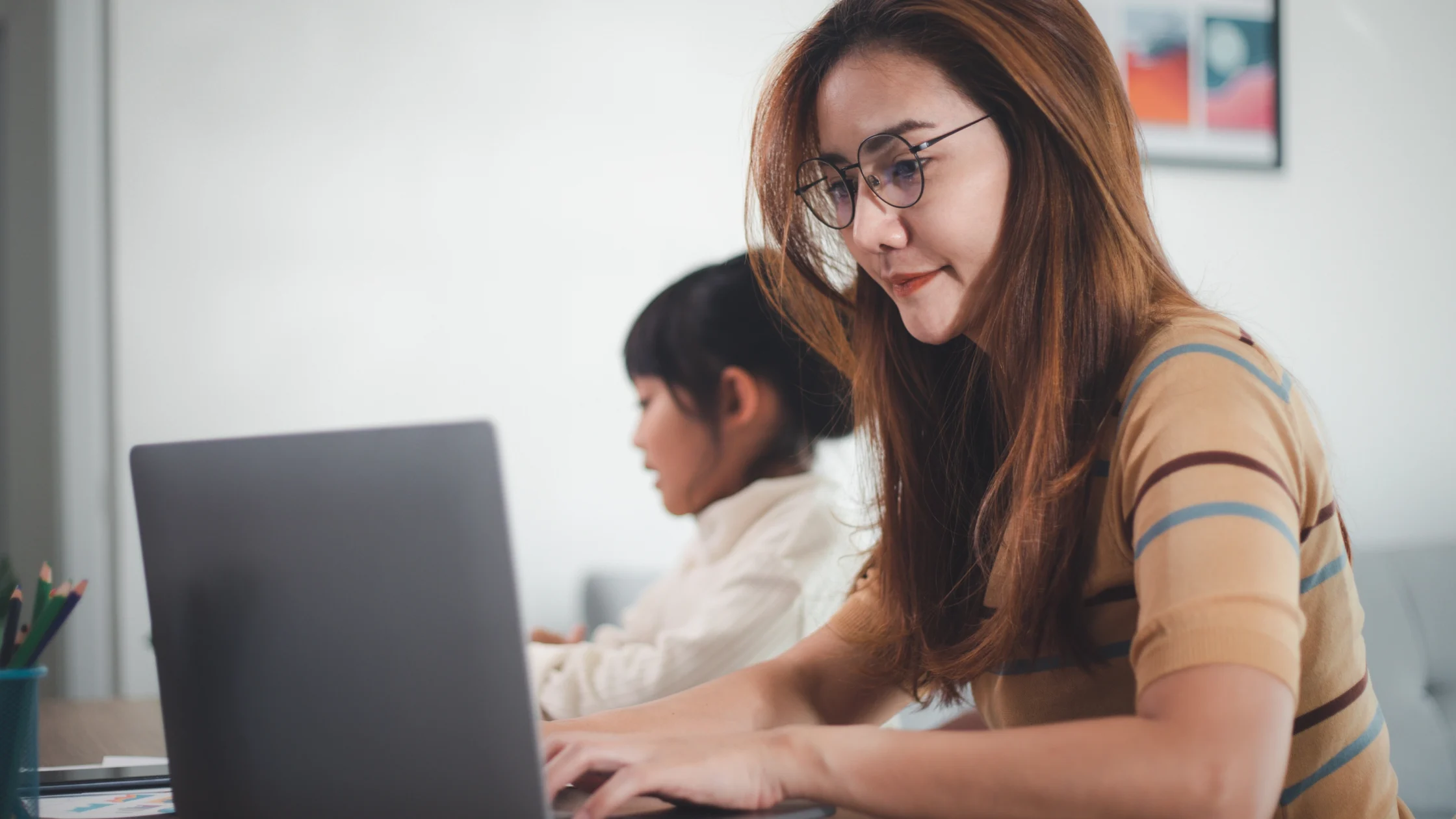 Woman with long hair and glasses typing on a laptop at a desk in an office, another person blurred in the background