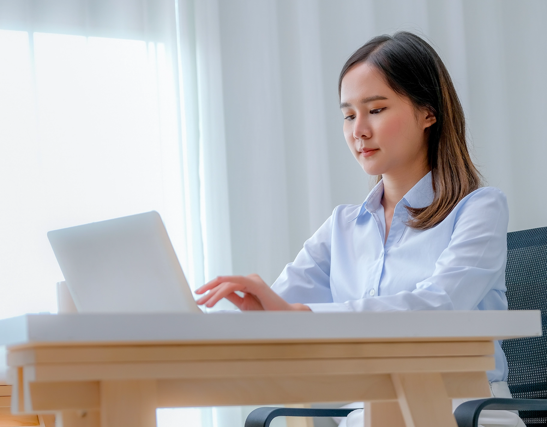 Woman in a light blue shirt typing on a laptop at a wooden desk in a bright office setting with sheer curtains behind her.