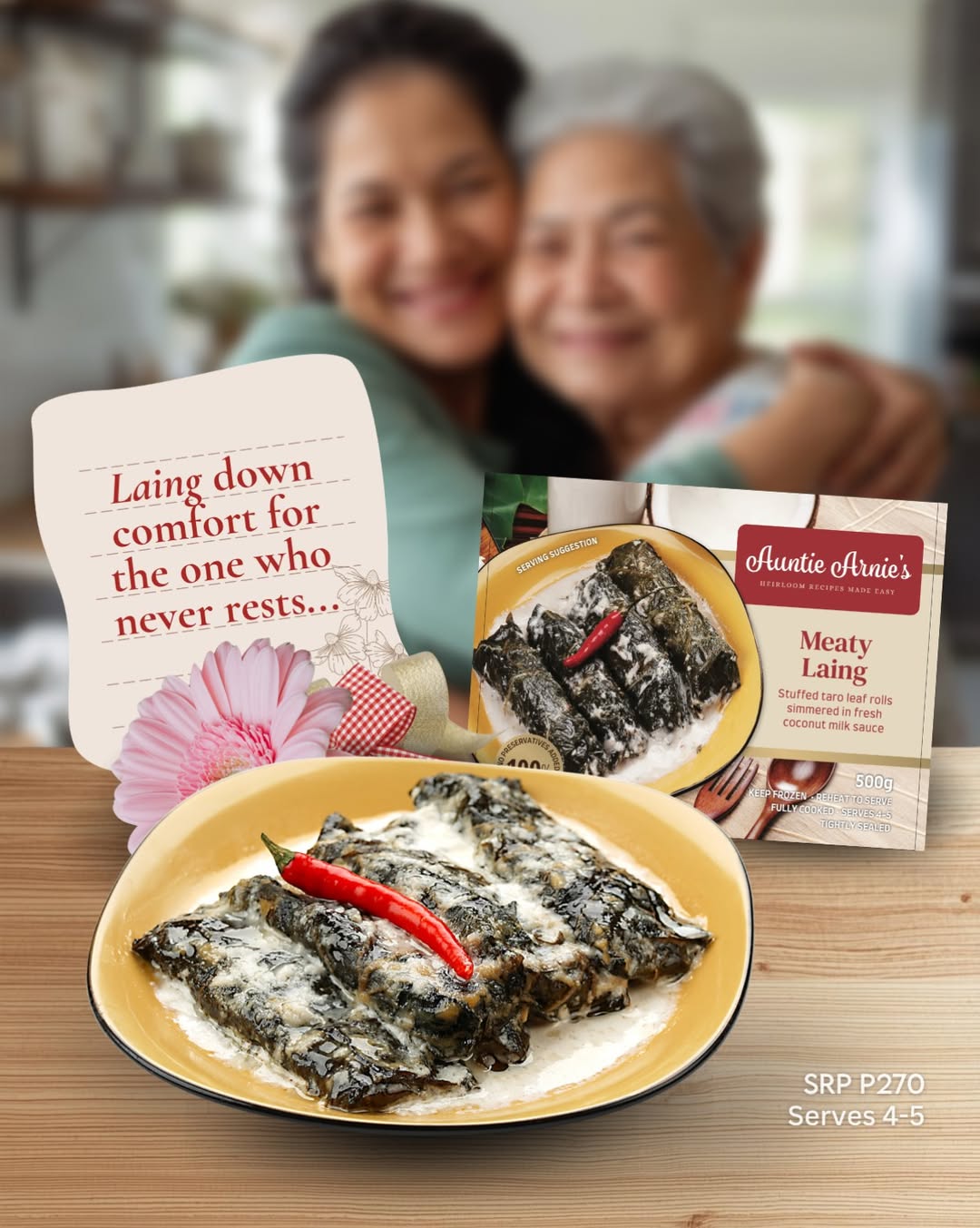 Two smiling people hug in the blurred background while a ready-made Auntie Arnie's Meaty Laing and a plated taro leaf dish sit on a wooden table in the foreground.