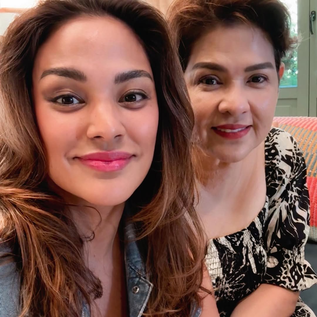 Two women take a close-up selfie indoors, smiling at the camera on a striped orange couch; one with long brown hair and denim jacket, the other with short dark hair in a black-and-white patterned top.