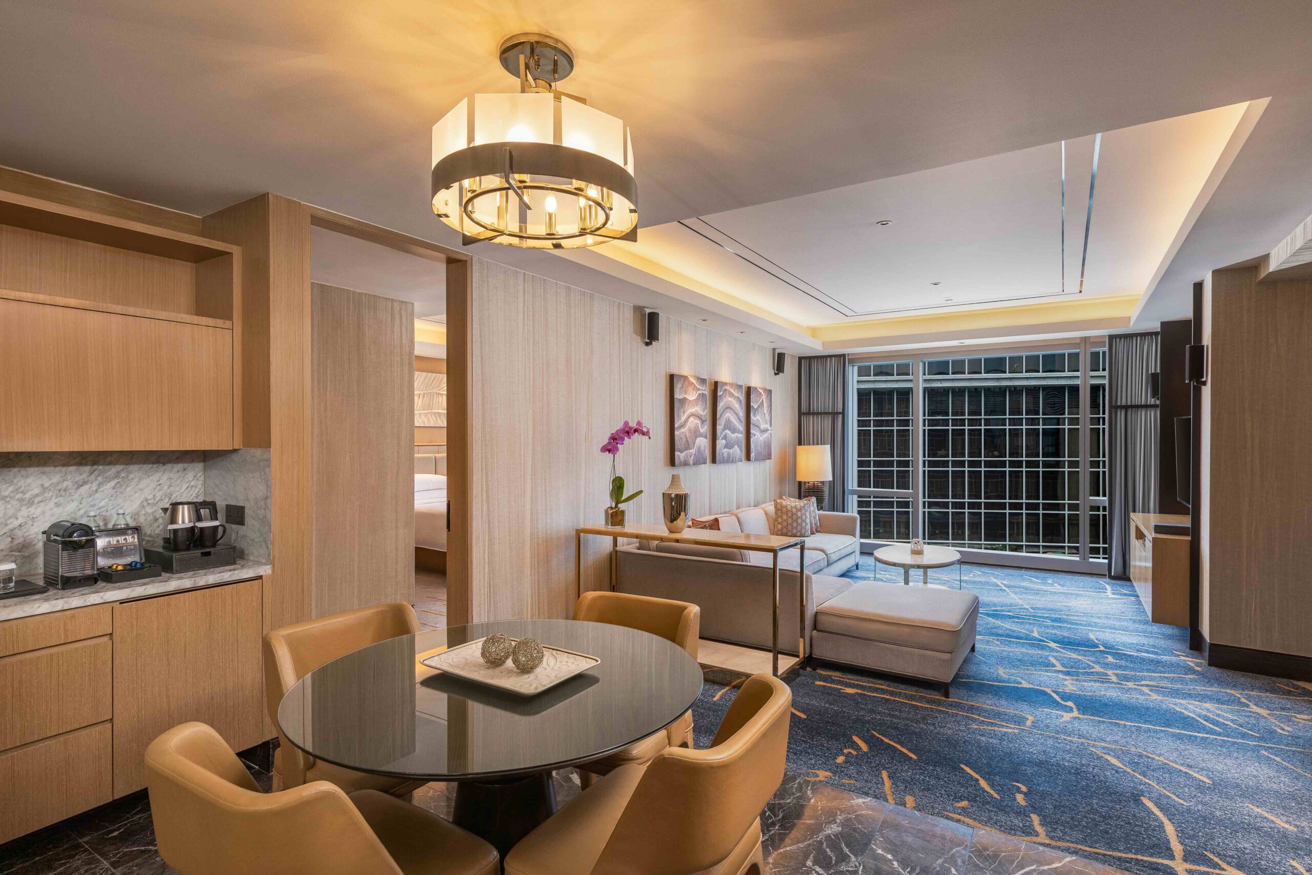 Open-concept hotel suite with a round glass dining table, tan chairs, and a living area by large windows on the right.