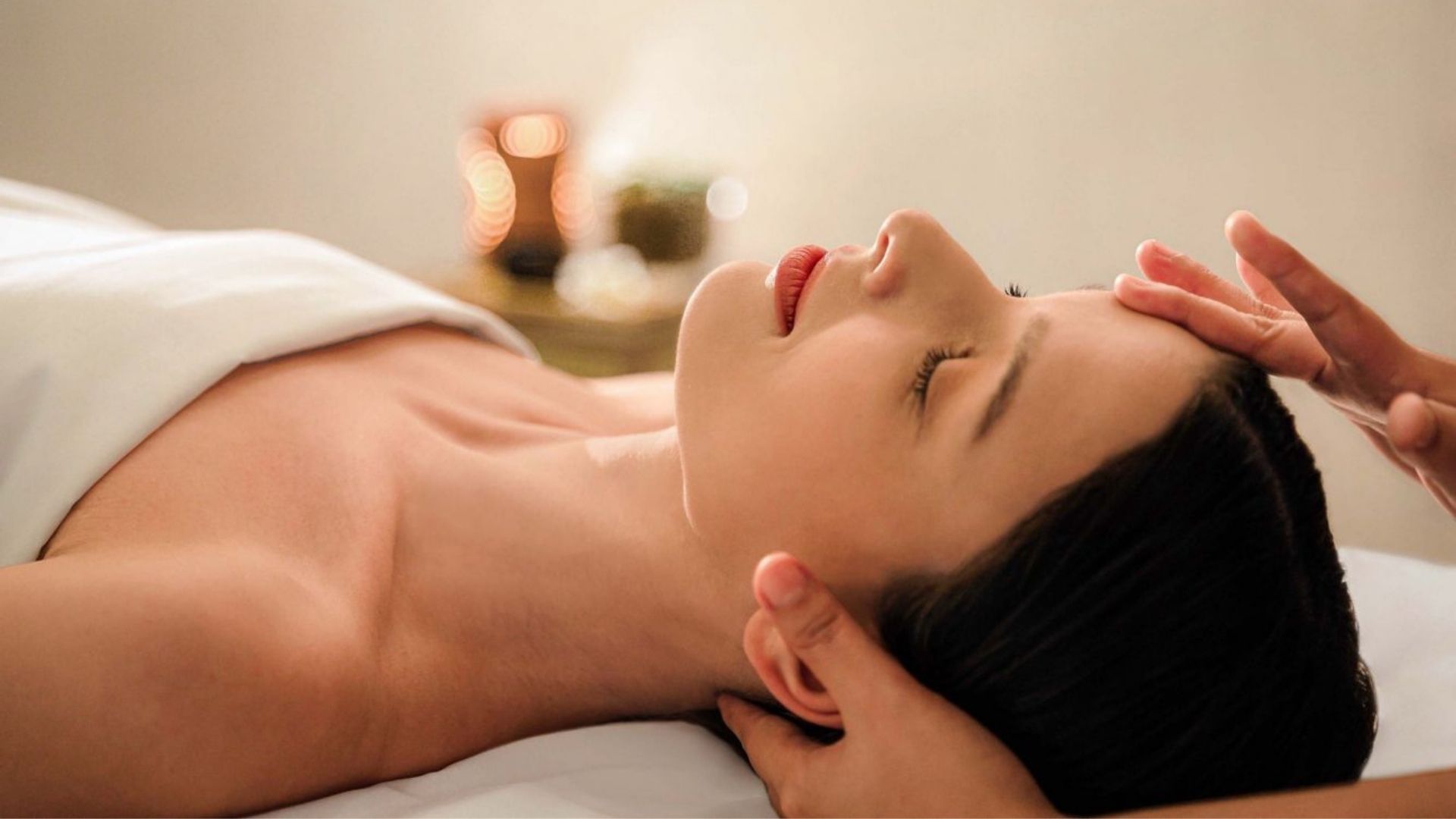 Woman lying down with eyes closed during a facial treatment as a therapist supports her head with both hands.