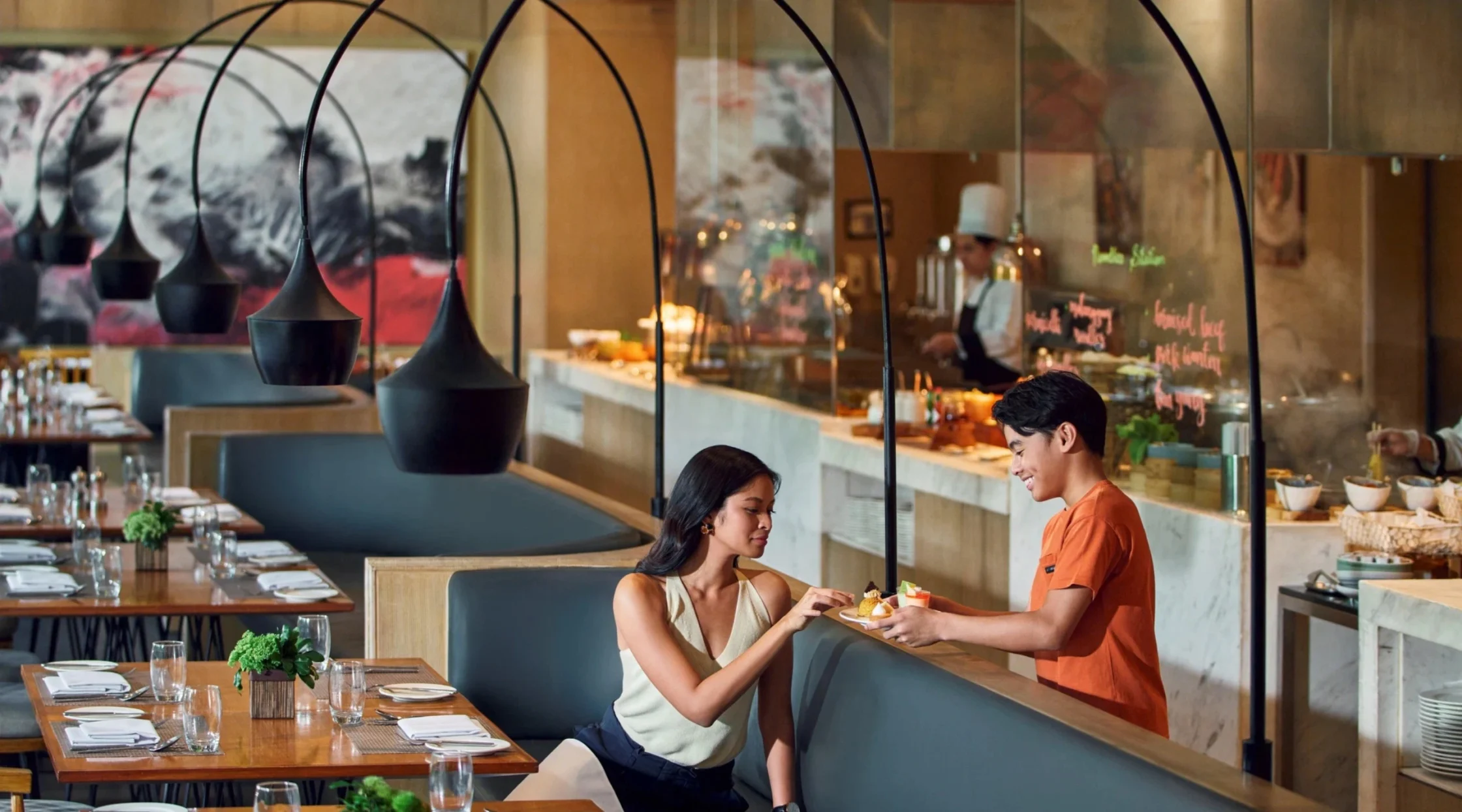 Couple sharing dessert at a table in a modern open-kitchen restaurant; a waiter in orange serves nearby.